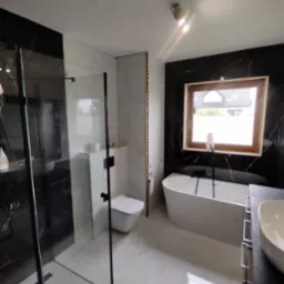 Nowoczesna łazienka z kabiną prysznicową typu walk-in, wiszącą toaletą, wolnostojącą wanną i umywalką nablatową, wykończona czarnym marmurem i białymi płytkami imitującymi marmur z delikatnym...