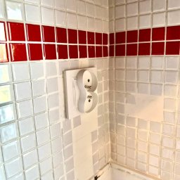 EIM - Paweł Mróz - Białe gniazdko elektryczne podwójne z wystającymi wtykami na ścianie z białych płytek z czerwoną linią dekoracyjną.