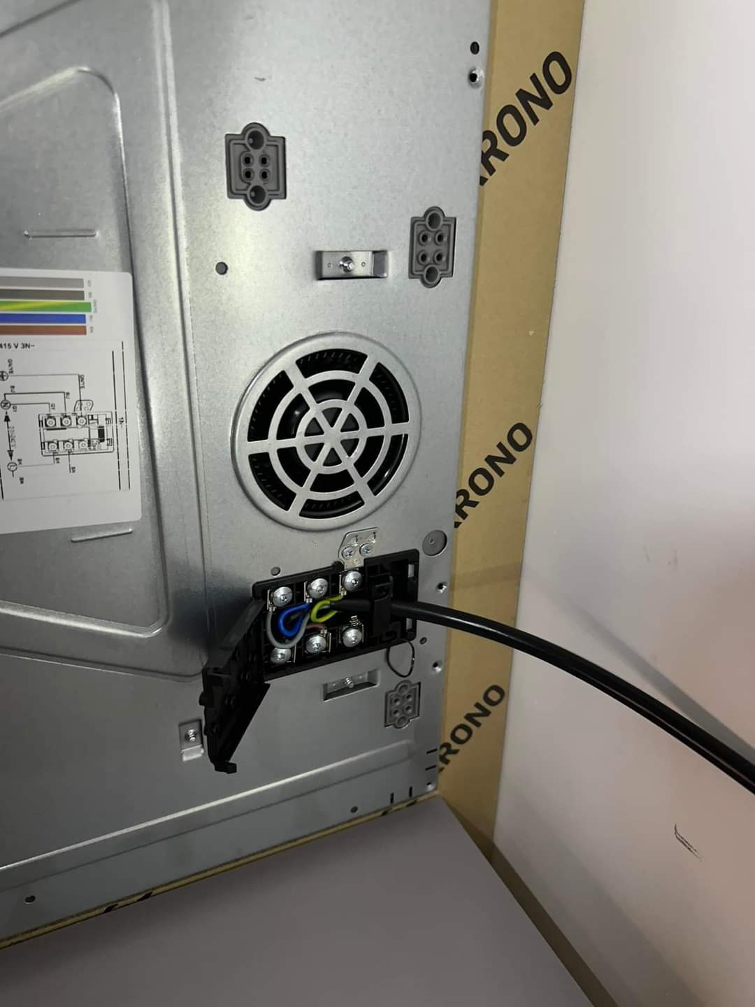 Widok z tyłu płyty indukcyjnej z podłączonym kablem zasilającym do listwy zaciskowej; widoczne przewody: niebieski, brązowy, żółto-zielony.