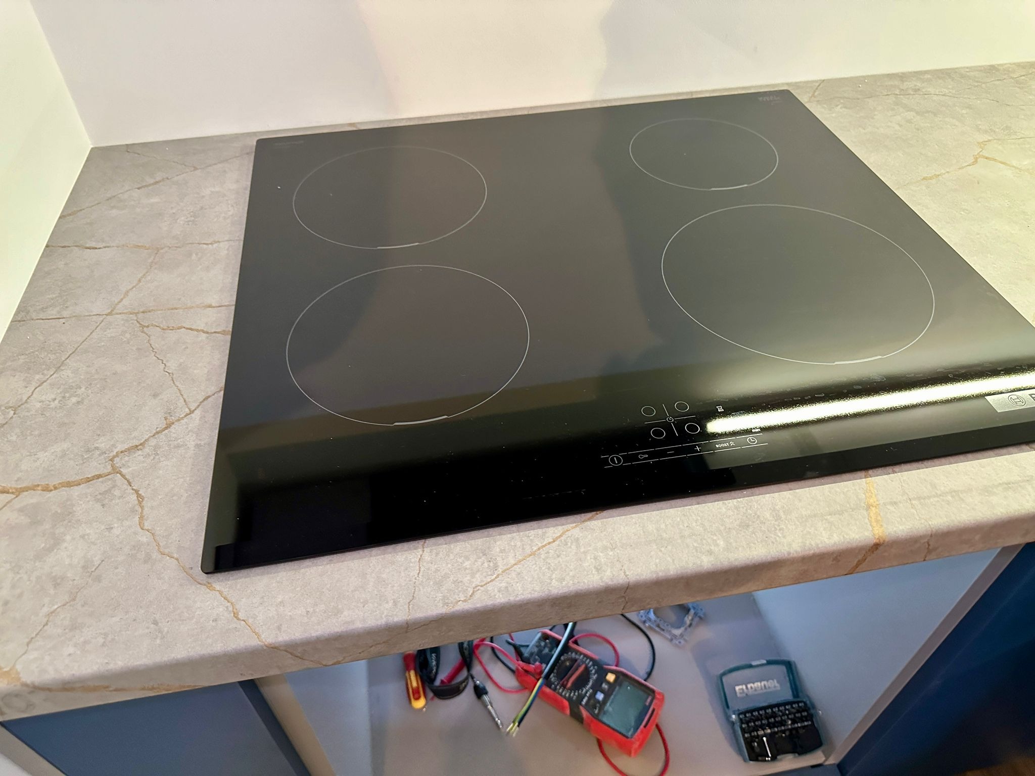 Czarna płyta indukcyjna czteropalnikowa wbudowana w blat o wzorze marmuru, widoczne narzędzia elektryczne pod spodem.