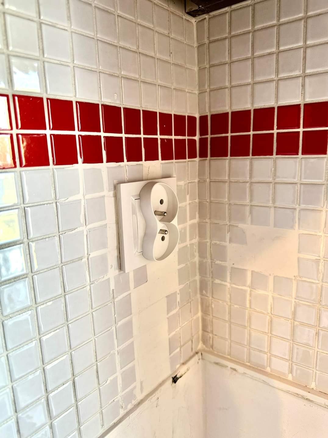 Białe gniazdko elektryczne podwójne z wystającymi wtykami na ścianie z białych płytek z czerwoną linią dekoracyjną.