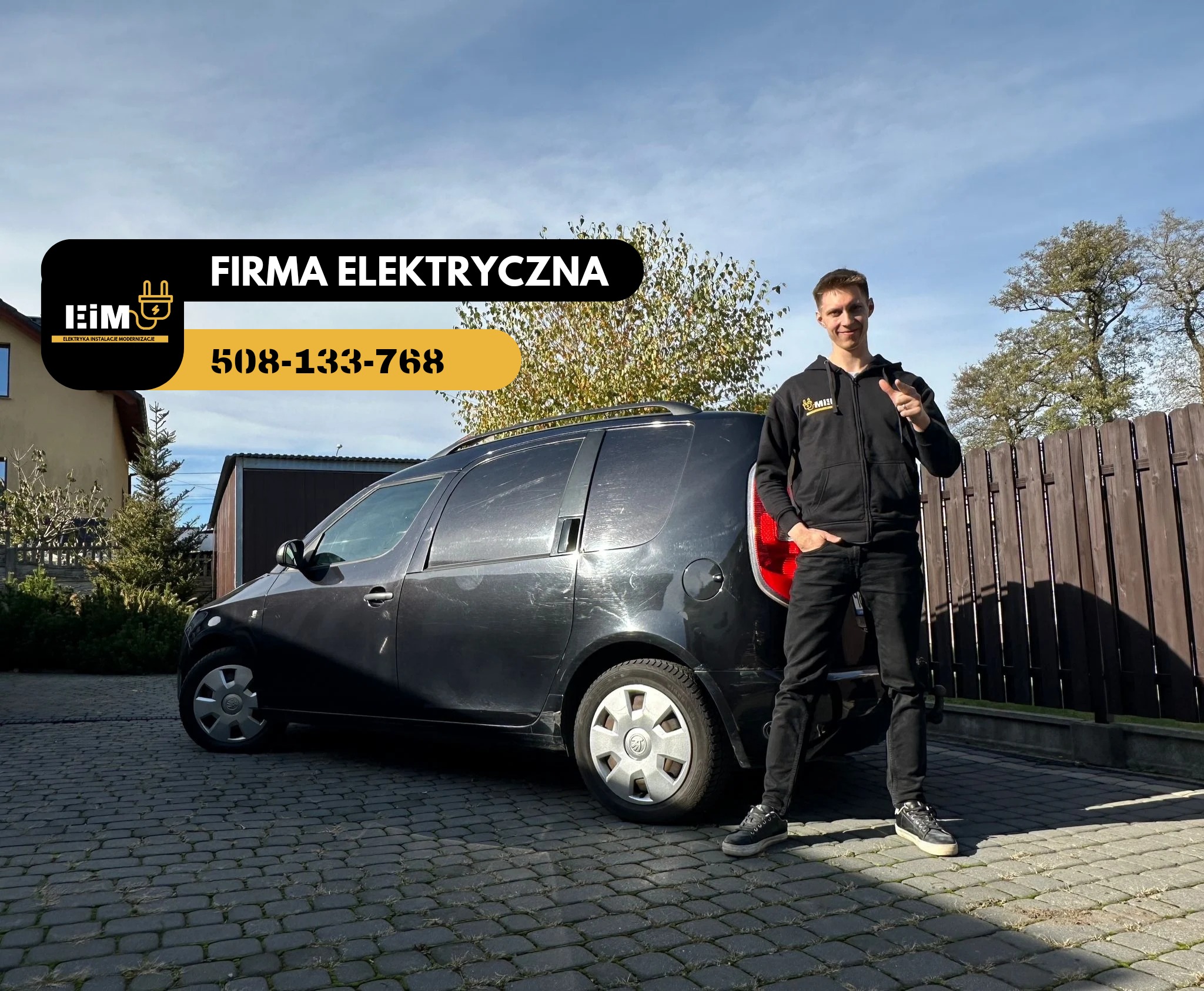 Elektryk w czarnej bluzie z logo firmy pozuje przed czarnym samochodem dostawczym z logo i numerem telefonu firmy na tle domu i ogrodzenia.