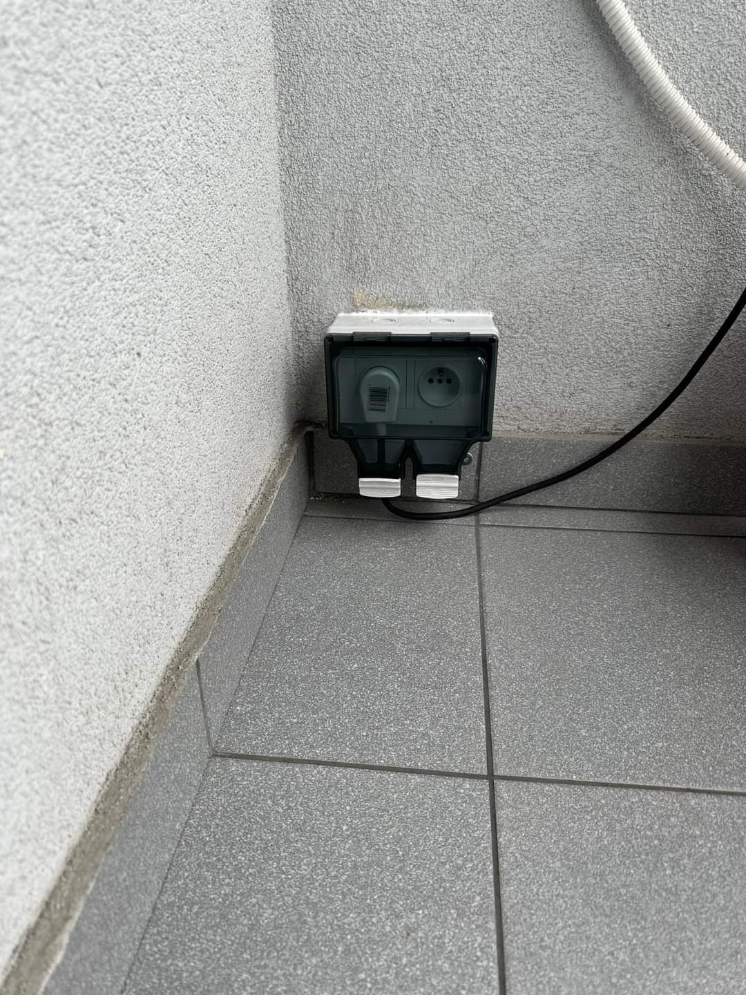 Zewnętrzne gniazdo elektryczne podwójne z klapką, zamontowane na szarej ścianie, widok z bliska na szarej podłodze z płytek.