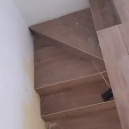 Układanie drewnopodobnych paneli na schodach, widoczne niedociągnięcia przy ścianie, narzędzie pomiarowe na schodku.