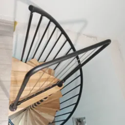 Spiralne schody z jasnego drewna z czarną metalową balustradą, widok z góry, fragment ściany z widocznymi instalacjami elektrycznymi.