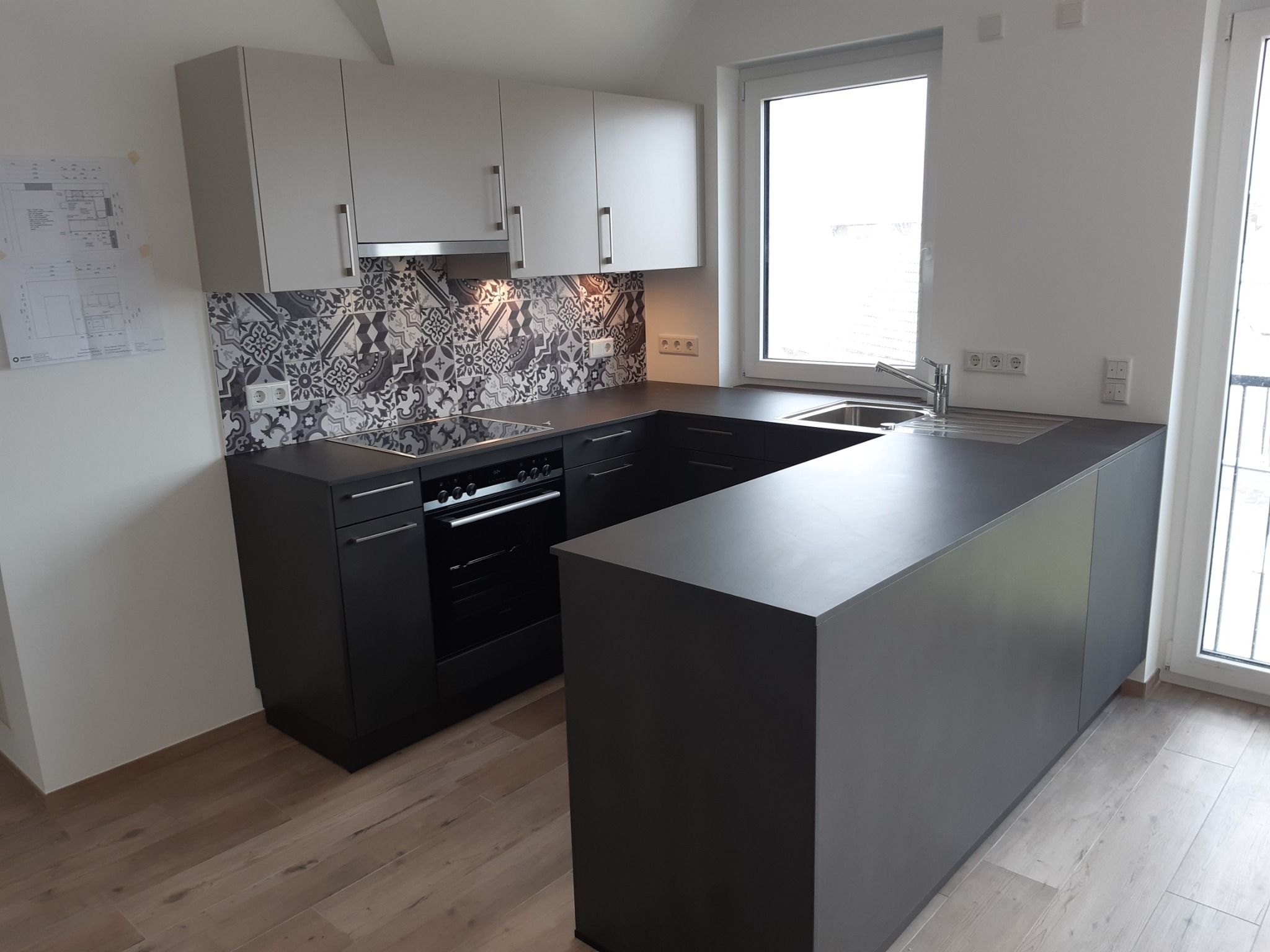 Nowoczesna kuchnia z szarymi szafkami górnymi i czarnymi dolnymi, wzorzysta mozaika nad blatem, drewnopodobna podłoga, okno z widokiem na zewnątrz.