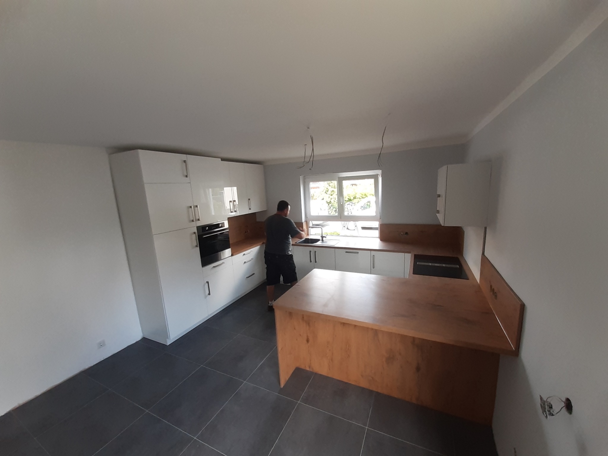 Montaż białych, lakierowanych mebli kuchennych z drewnianym blatem i wyspą; widoczne niedokończone podłączenia elektryczne.