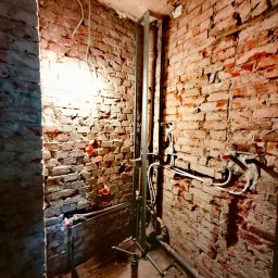 Usługi Remontowo Budowlane - Surowa ściana z czerwonej cegły z widocznymi przewodami elektrycznymi i instalacją hydrauliczną w trakcie montażu, oświetlona pojedynczą żarówką.