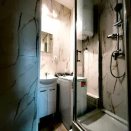 Wykończona łazienka z kabiną prysznicową, umywalką z szafką, pralką i bojlerem, wyłożona płytkami imitującymi marmur z brązowymi żyłami, oświetlona punktowo.