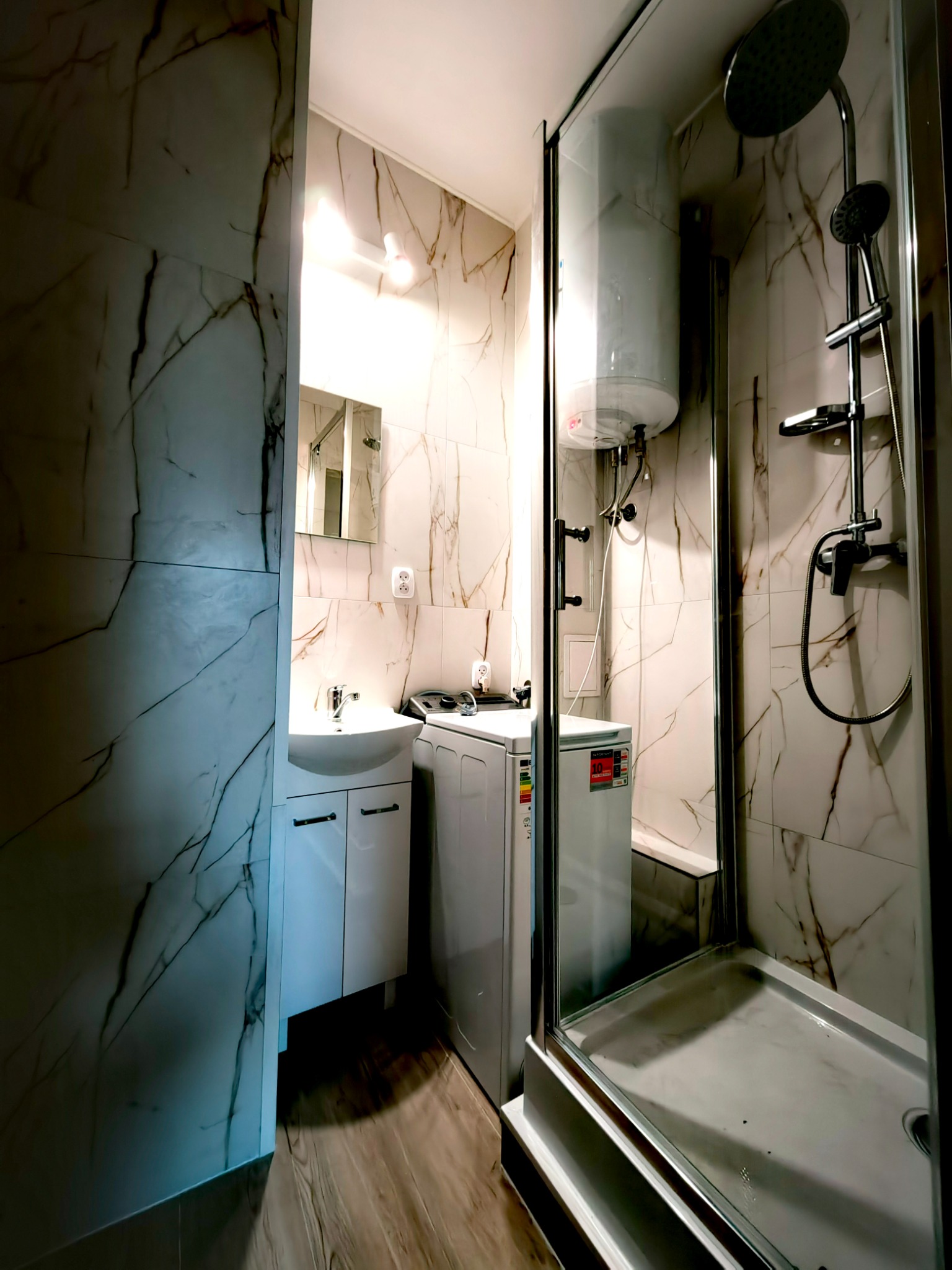 Wykończona łazienka z kabiną prysznicową, umywalką z szafką, pralką i bojlerem, wyłożona płytkami imitującymi marmur z brązowymi żyłami, oświetlona punktowo.