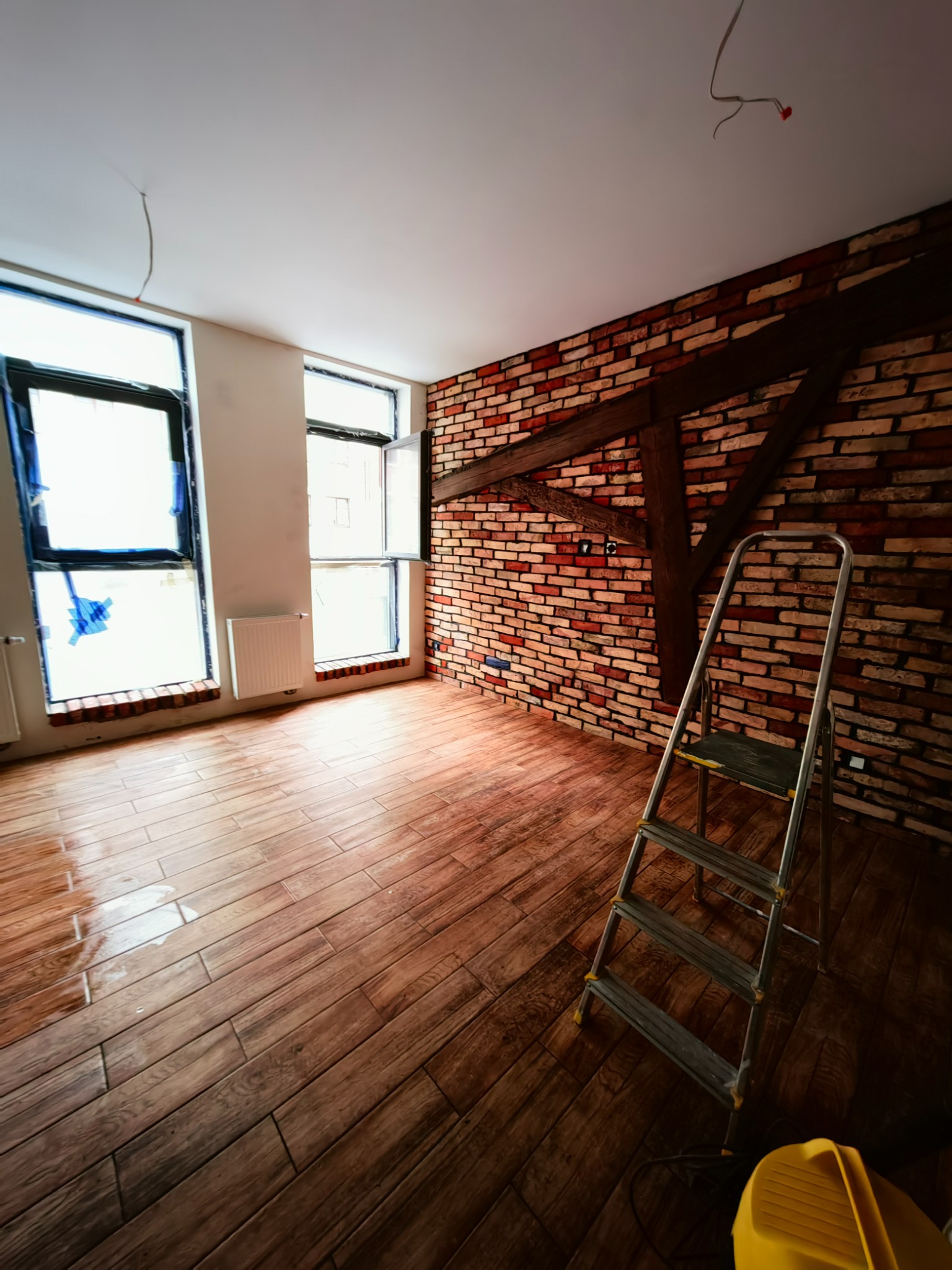 Wnętrze pokoju po remoncie: brązowe panele podłogowe imitujące drewno, ściana z cegły, drewniana belka, okna zabezpieczone folią, drabina aluminiowa i żółty odkurzacz przemysłowy.
