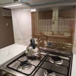 Krzysztof Sosinka - Nowoczesna kuchnia: płyta gazowa ze srebrnym czajnikiem, marmurowy blat, szklany panel nad płytą, okap Candy i oświetlenie LED. Minimalistyczny design.