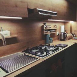 Krzysztof Sosinka - Nowoczesna kuchnia z drewnianymi frontami, oświetleniem LED podszafkowym, blatem roboczym i wyposażeniem: zlew, bateria, płyta gazowa, okap, półki na przyprawy.