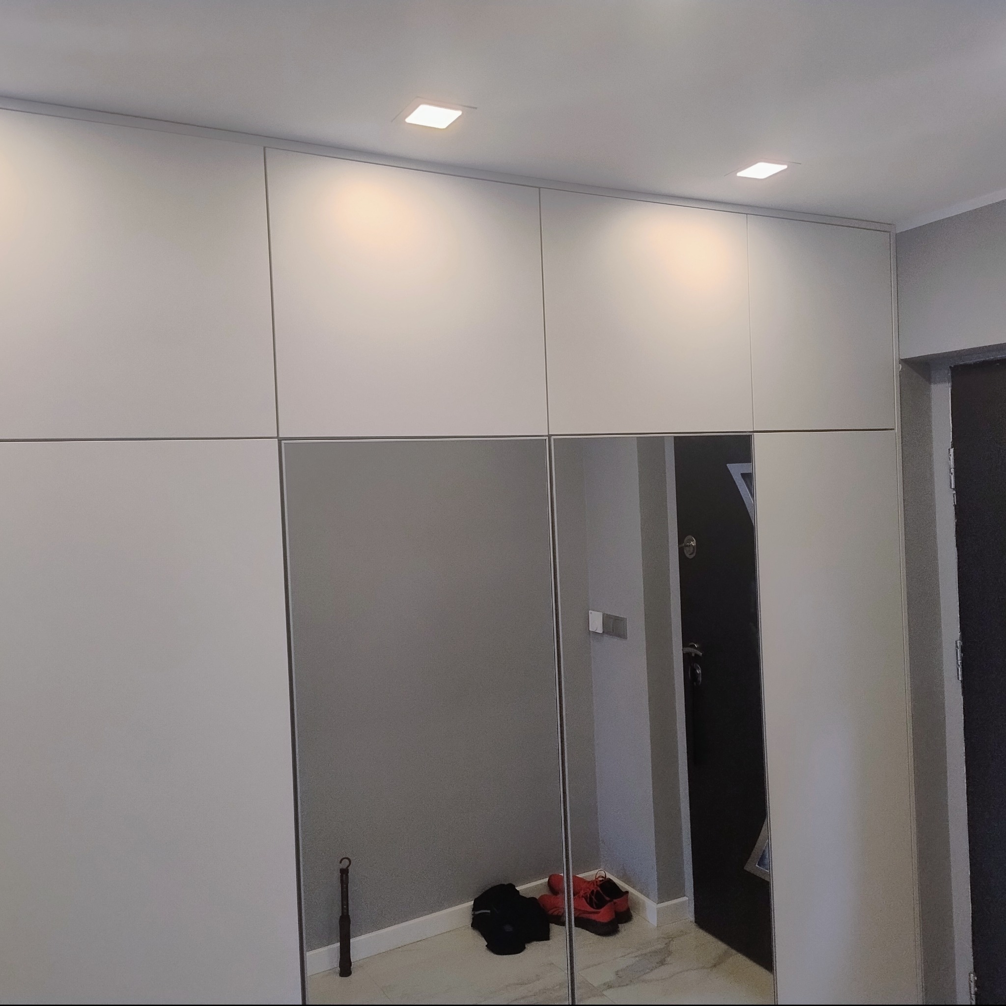 Biała szafa wnękowa z lustrzanymi drzwiami, oświetleniem punktowym na suficie, odbijająca fragment przedpokoju z butami i parasolem. Minimalistyczny design.