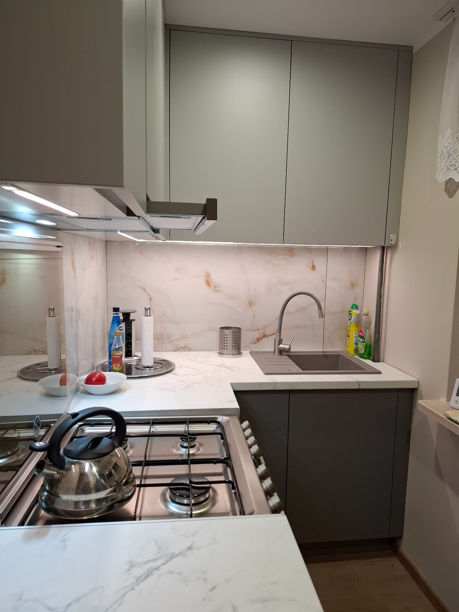 Nowoczesna kuchnia z szarymi szafkami, marmurowym blatem i podświetleniem LED. Na blacie czajnik, zlew z baterią i akcesoria kuchenne. Minimalistyczny design.