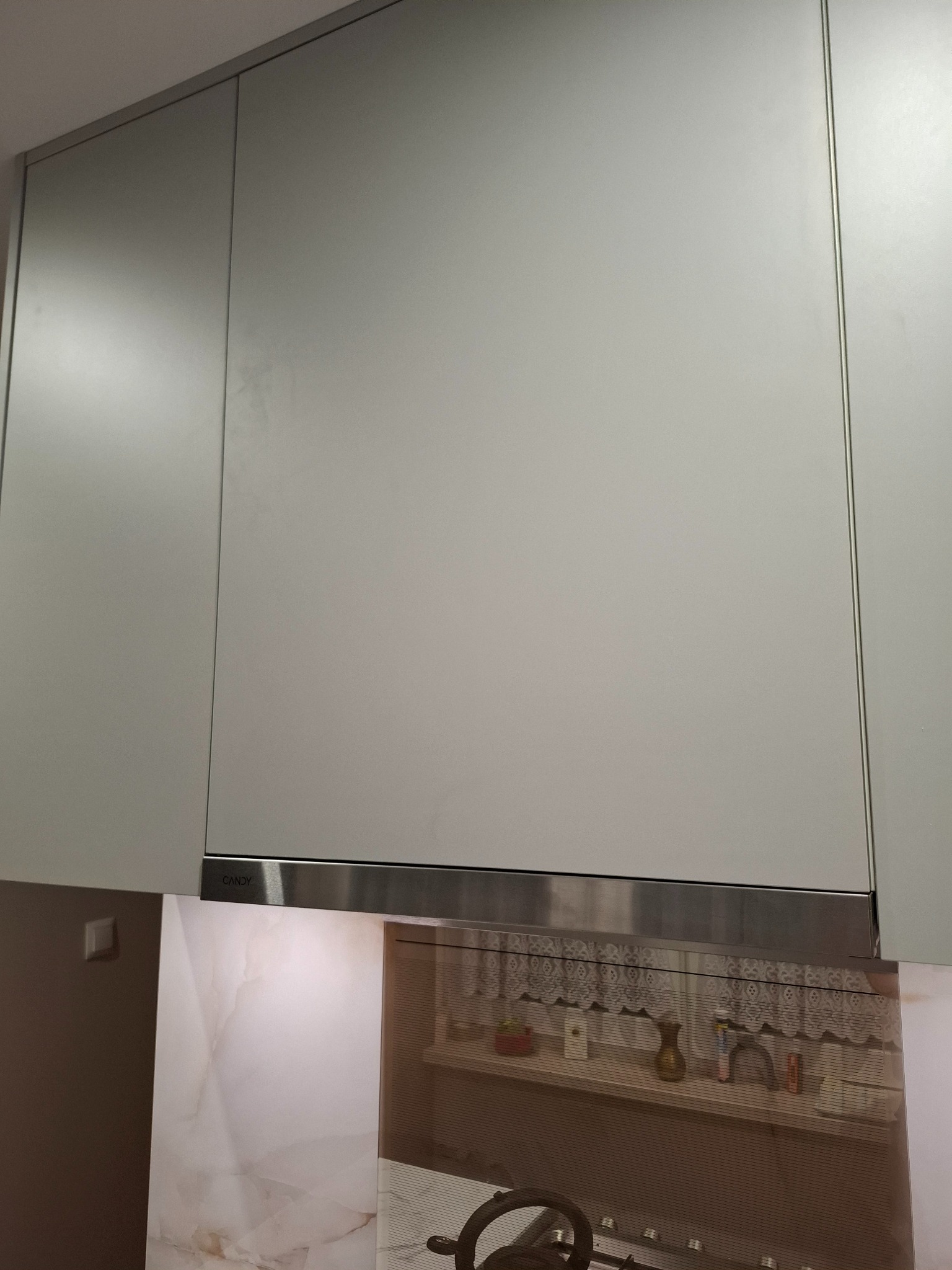 Jasnoszare szafki kuchenne podwieszane z okapem Candy, widoczne detale wykończenia i fragment blatu z kuchenką. Minimalistyczny design.