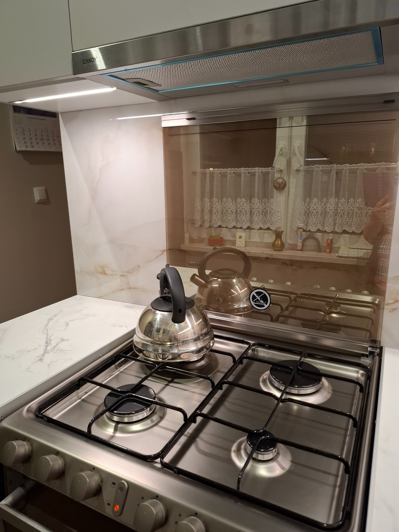 Nowoczesna kuchnia: płyta gazowa ze srebrnym czajnikiem, marmurowy blat, szklany panel nad płytą, okap Candy i oświetlenie LED. Minimalistyczny design.
