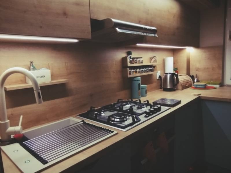 Nowoczesna kuchnia z drewnianymi frontami, oświetleniem LED podszafkowym, blatem roboczym i wyposażeniem: zlew, bateria, płyta gazowa, okap, półki na przyprawy.