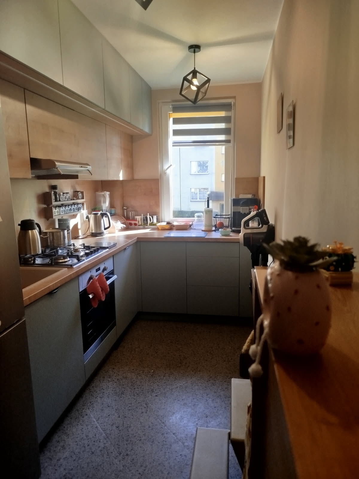 Kompaktowa kuchnia w bloku z szarymi frontami i drewnianymi blatami, oświetlona nowoczesną lampą wiszącą. Widok na okno z roletami i budynek na zewnątrz.