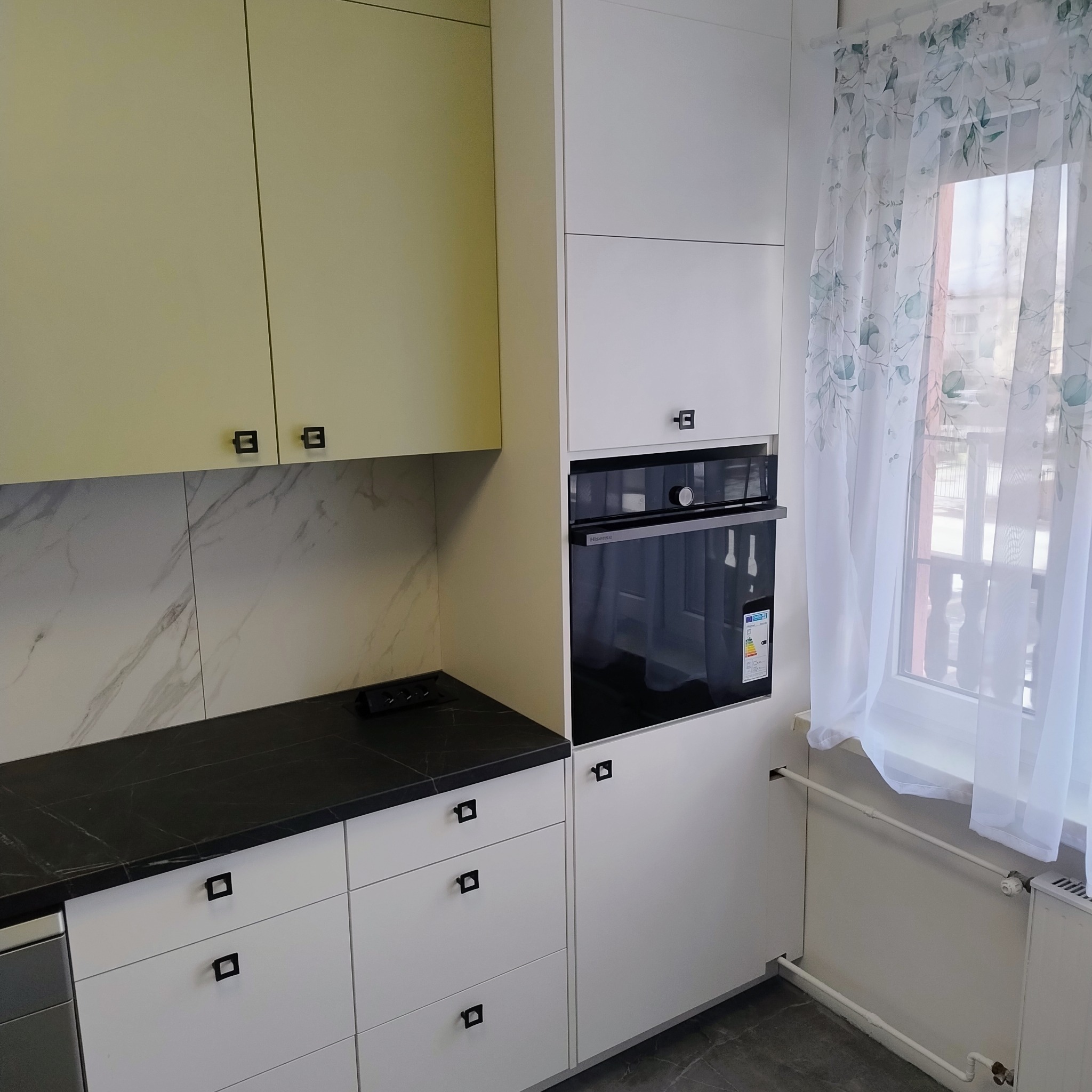 Jasna kuchnia z czarnym blatem i marmurową ścianą. Białe szafki z kwadratowymi uchwytami. Wbudowany piekarnik i okno z delikatną, wzorzystą firanką.