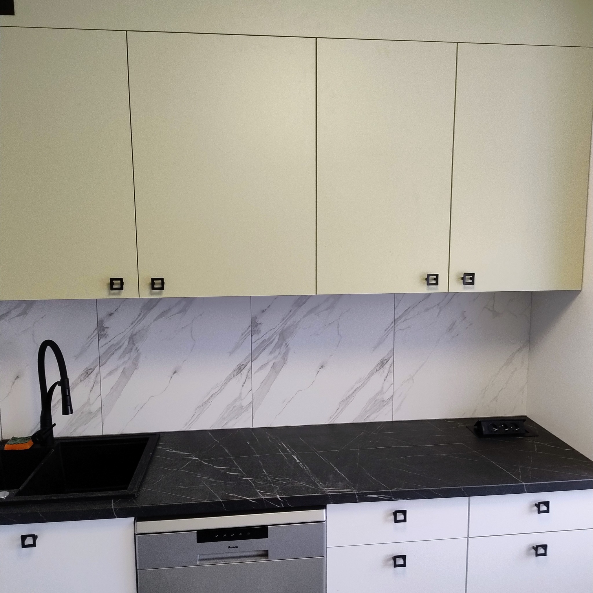 Jasne szafki wiszące z kwadratowymi uchwytami nad blatem z czarnego marmuru i marmurowymi płytkami. Widoczny zlew i zintegrowana z zabudową zmywarka.