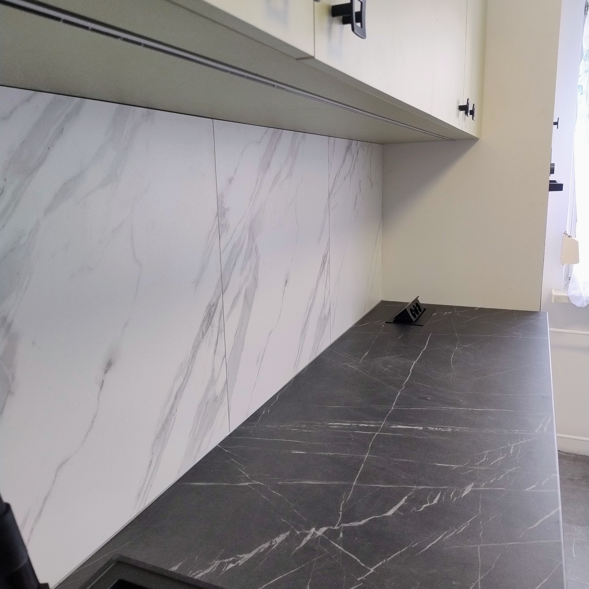 Fragment kuchni: blat z czarnego marmuru z białymi żyłami, ściana z białego marmuru, górne szafki z czarnymi uchwytami. Nowoczesny design.