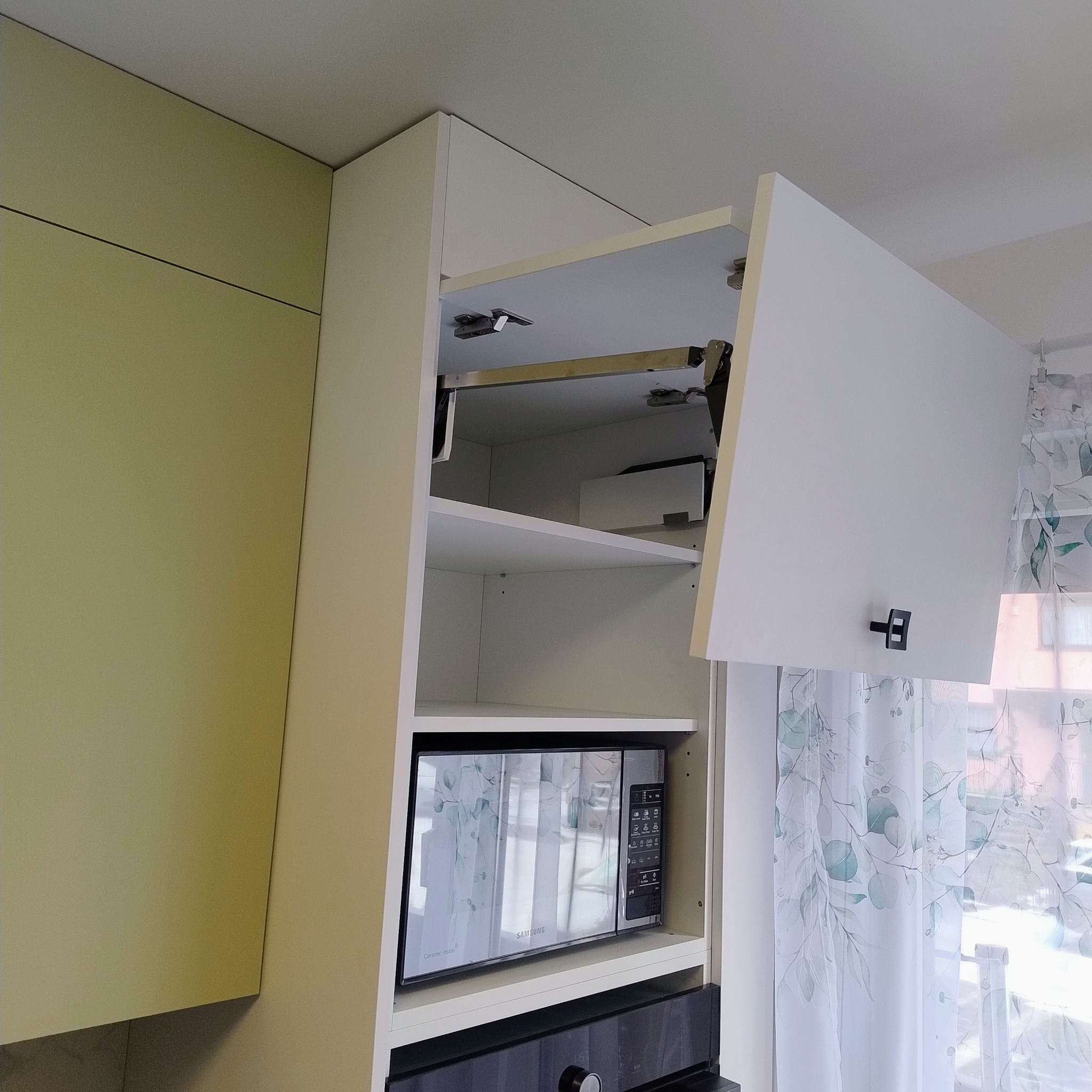 Biała szafka kuchenna z mechanizmem podnoszenia frontu, wewnątrz mikrofalówka Samsung. Widoczne jasne zasłony w tle. Ujęcie z dołu.