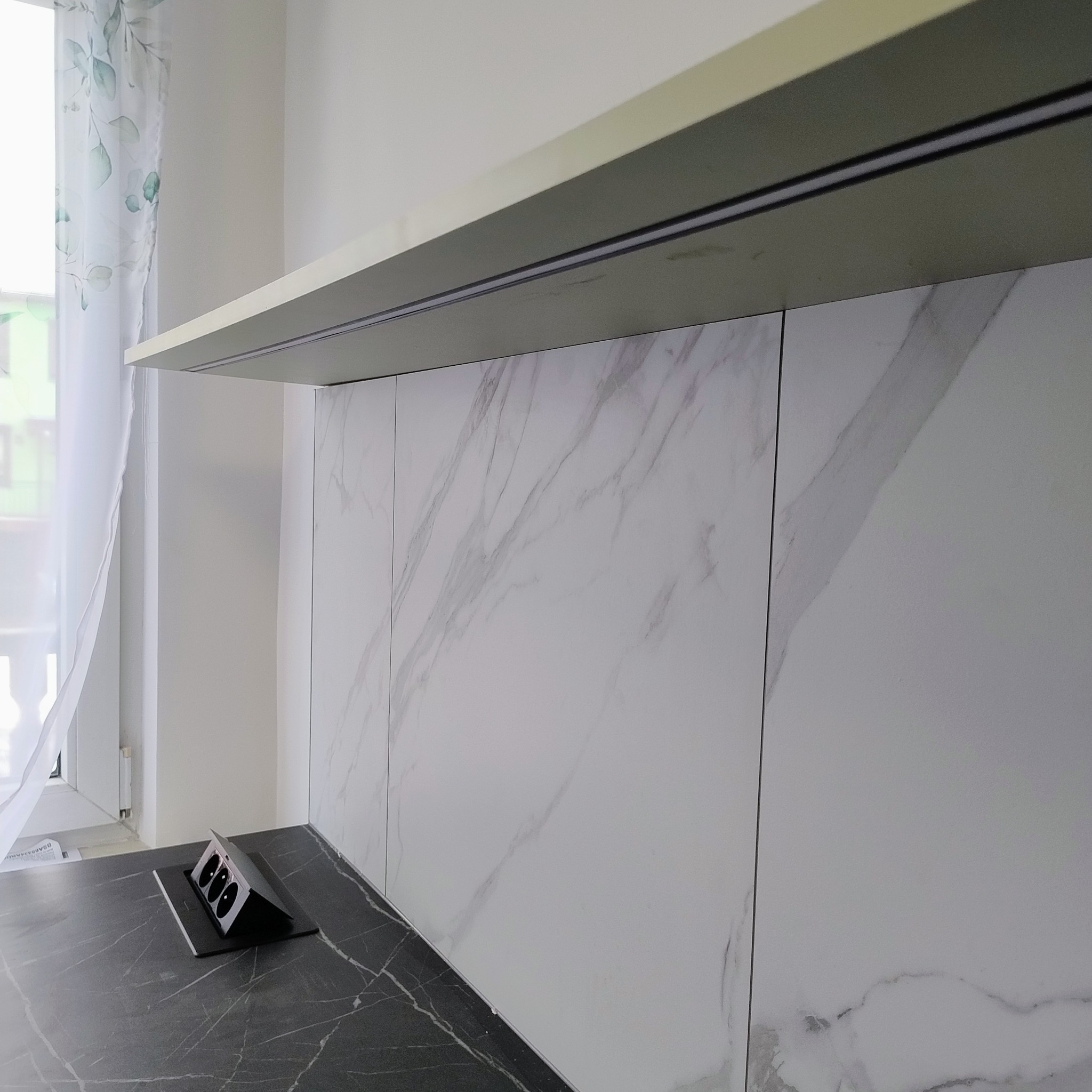Białe panele ścienne z marmurowym wzorem w nowoczesnej kuchni, widoczne gniazdka elektryczne w blacie i fragment okna z zasłoną w tle.