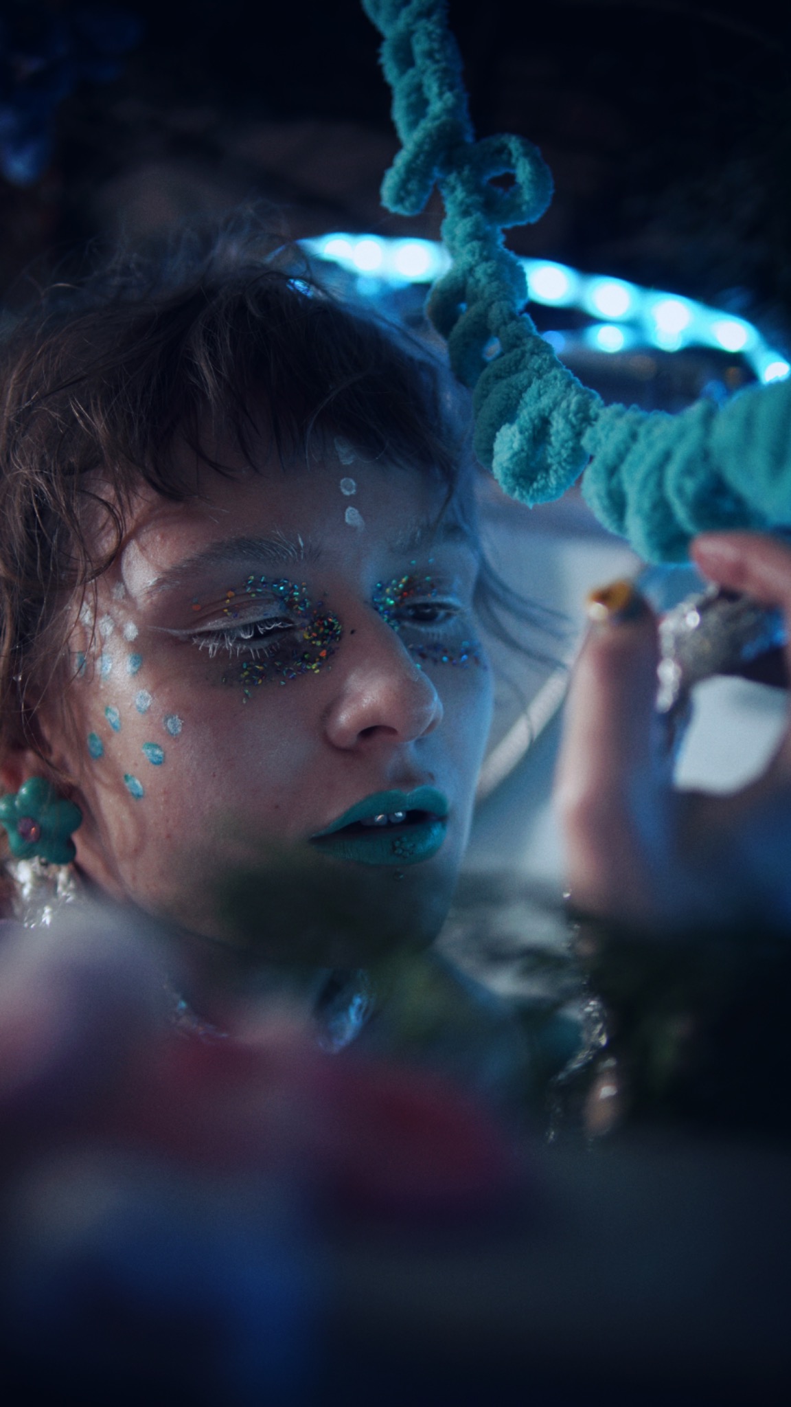 Portret osoby z artystycznym makijażem: brokat na powiekach, turkusowe usta, białe kropki na twarzy i kolczyk w wardze, oświetlona niebieskim światłem.