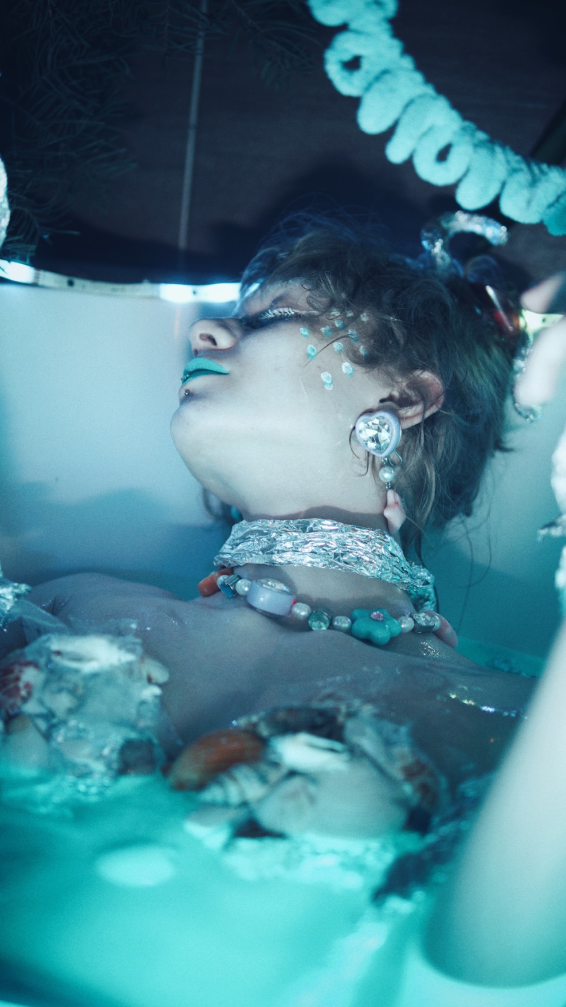 Portret kobiety leżącej w wodzie z turkusowymi ustami, srebrną folią na szyi, ozdobnymi kolczykami i makijażem z błyszczącymi kropkami na twarzy.