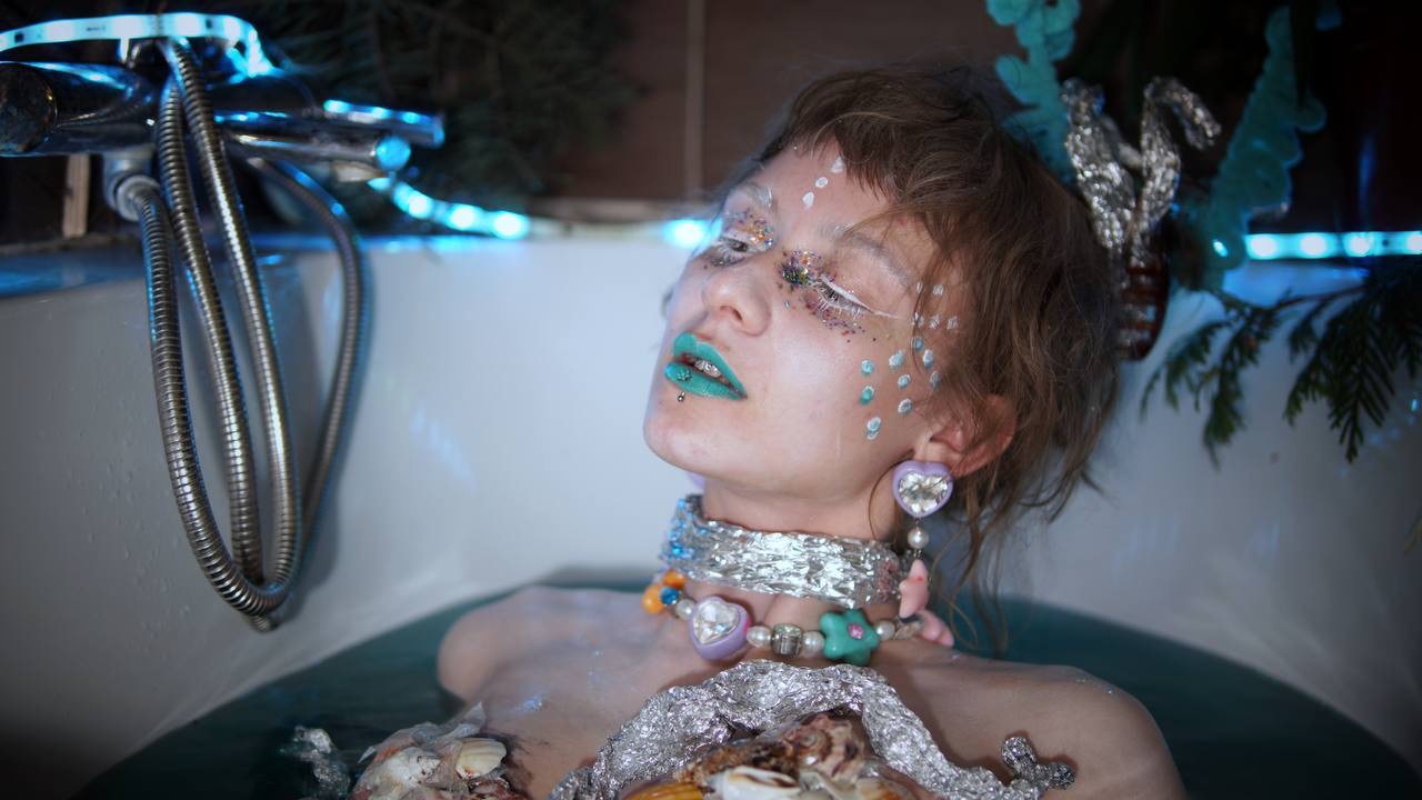 Artystyczna sesja w wannie: kobieta z turkusowymi ustami, brokatem na twarzy i muszlami, oświetlona niebieskim światłem.