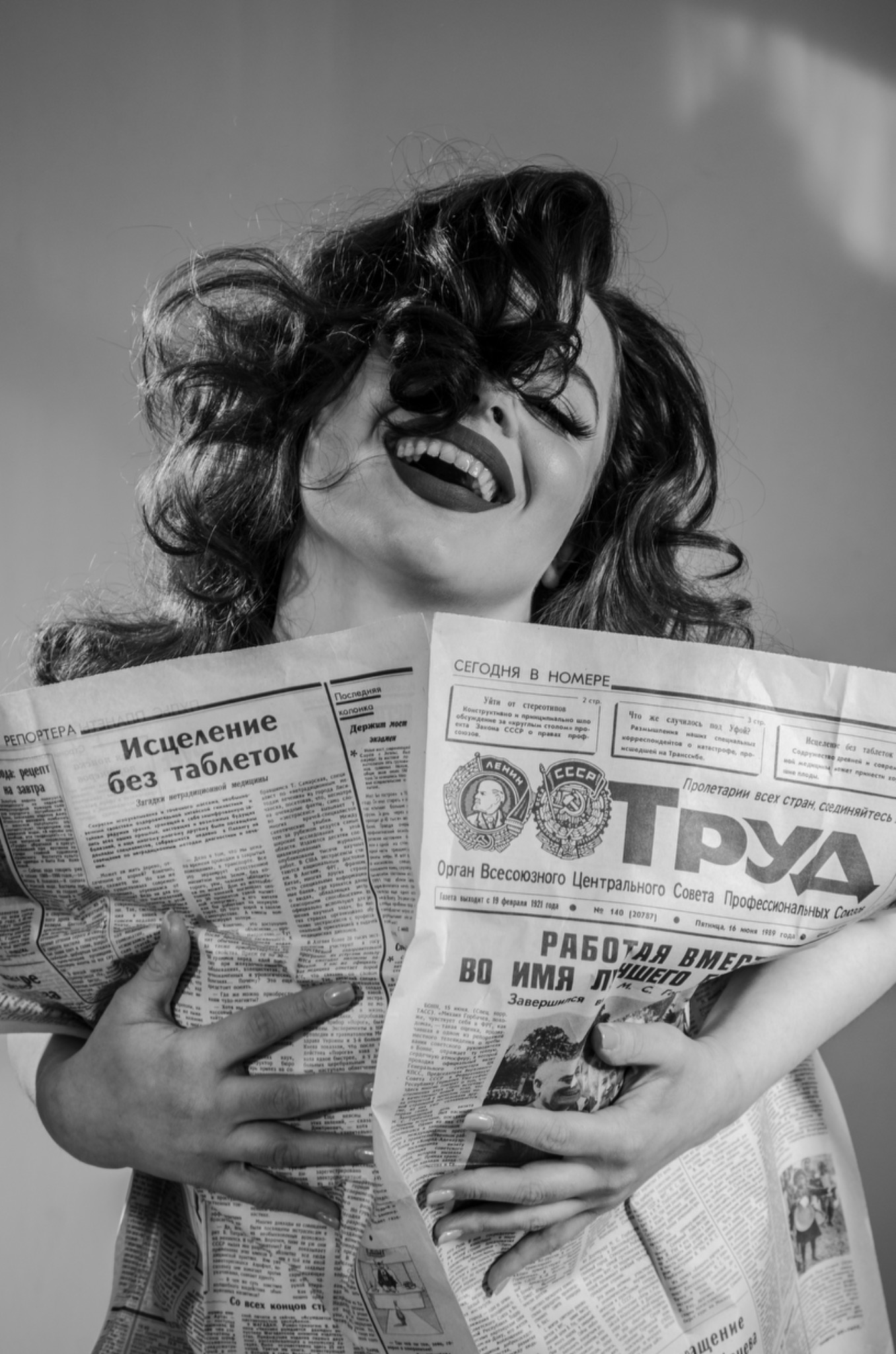 Czarno-białe zdjęcie uśmiechniętej kobiety z mocnym makijażem ust, trzymającej dwie rozłożone gazety z nagłówkami w języku rosyjskim, zasłaniające jej ciało.