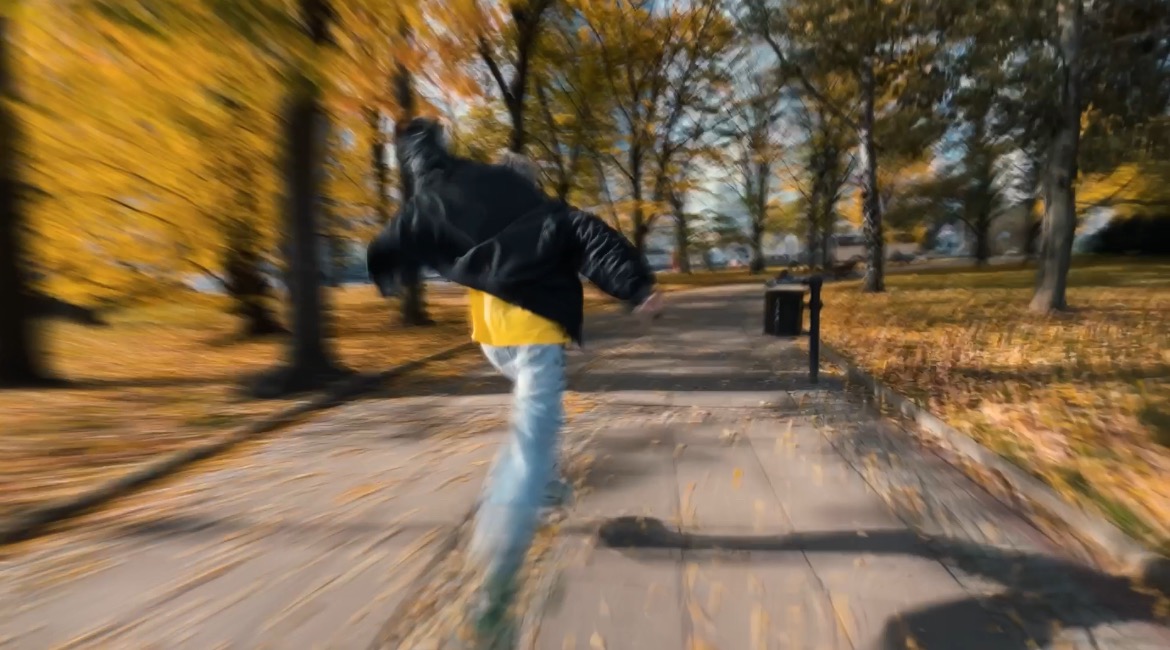 Osoba w ruchu, ubrana w czarną kurtkę i żółtą koszulkę, biegnie po chodniku w parku jesienią, efekt rozmycia ruchu podkreśla dynamikę ujęcia.