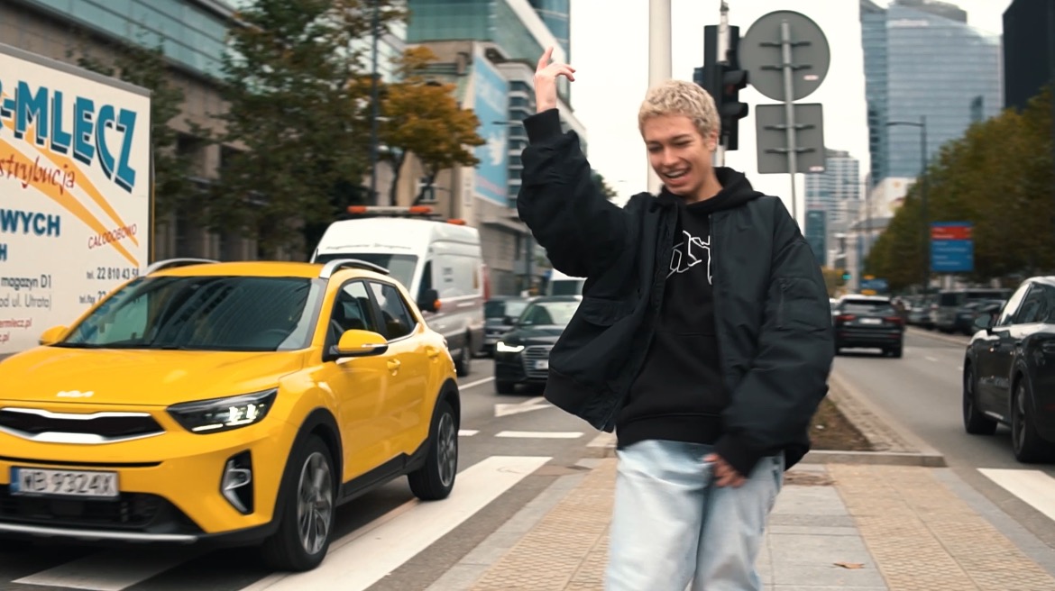 Młody mężczyzna z blond włosami tańczy na pasach na tle ulicy z żółtym samochodem i budynkami w Warszawie.