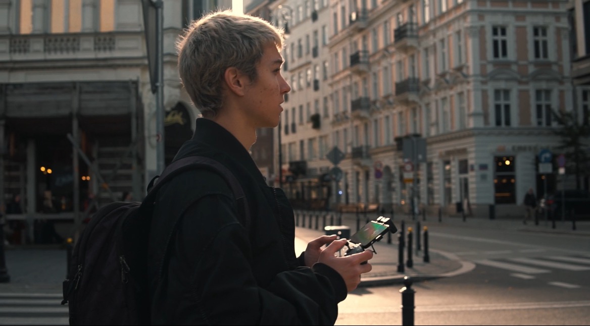 Młody mężczyzna z plecakiem, profil, trzyma kontroler drona na tle miejskiej ulicy z budynkami w stylu kamienic.