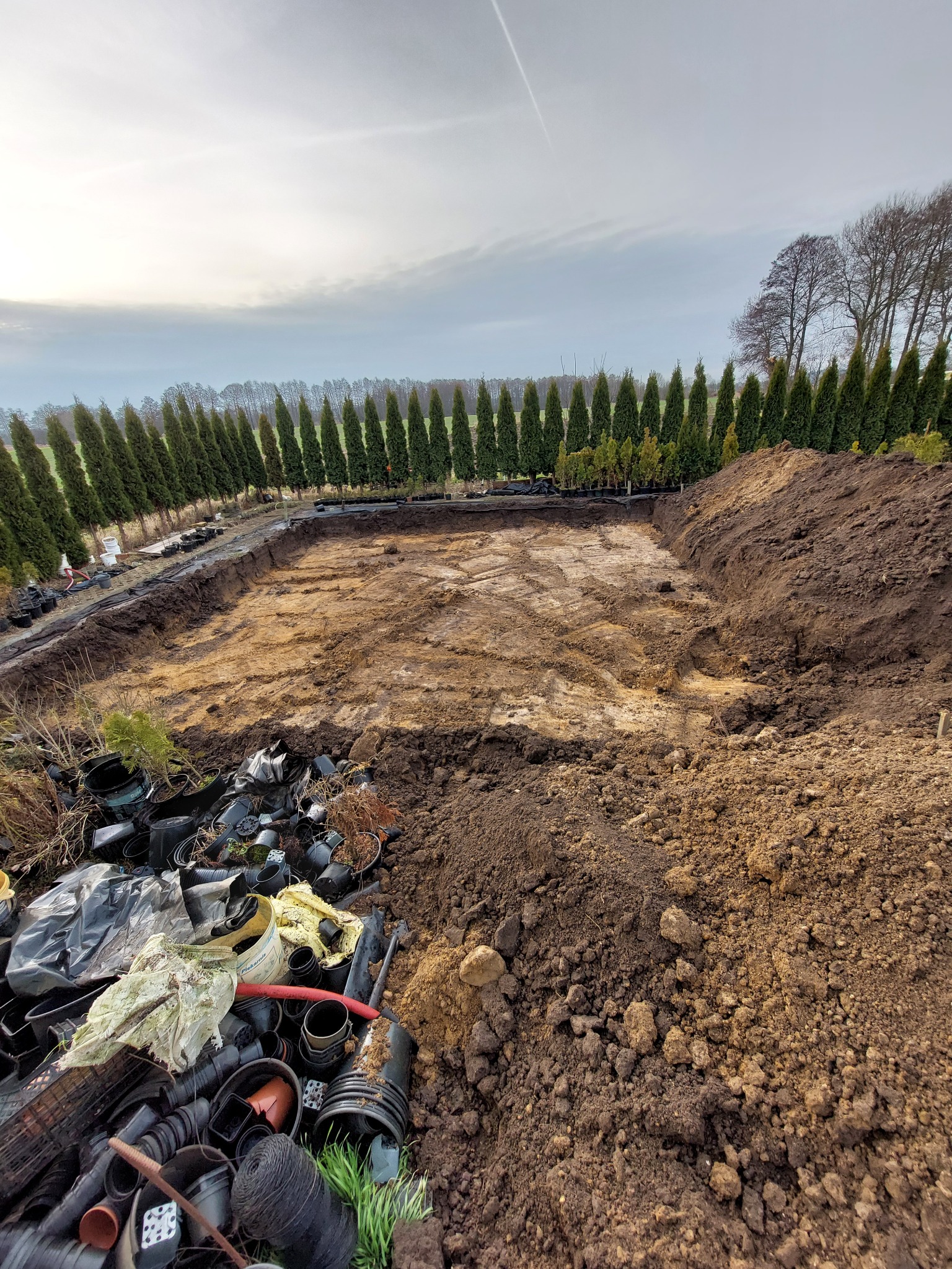 Wykonany wykop pod przyszły staw lub oczko wodne, obok sterta plastikowych doniczek i resztek roślin, w tle rząd tuj.