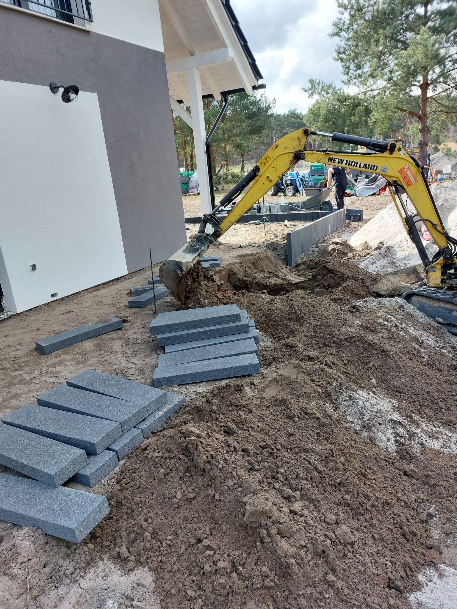 Żółta koparka New Holland podczas prac ziemnych przy budowie domu, widoczne ułożone szare obrzeża chodnikowe i wykopany rów.