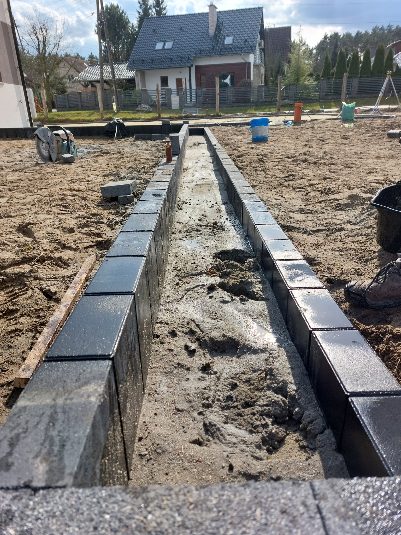 Wąski wykop z betonową podsypką, obramowany mokrymi, błyszczącymi kostkami w kolorze antracytu, widok z góry na plac budowy z domami w tle.