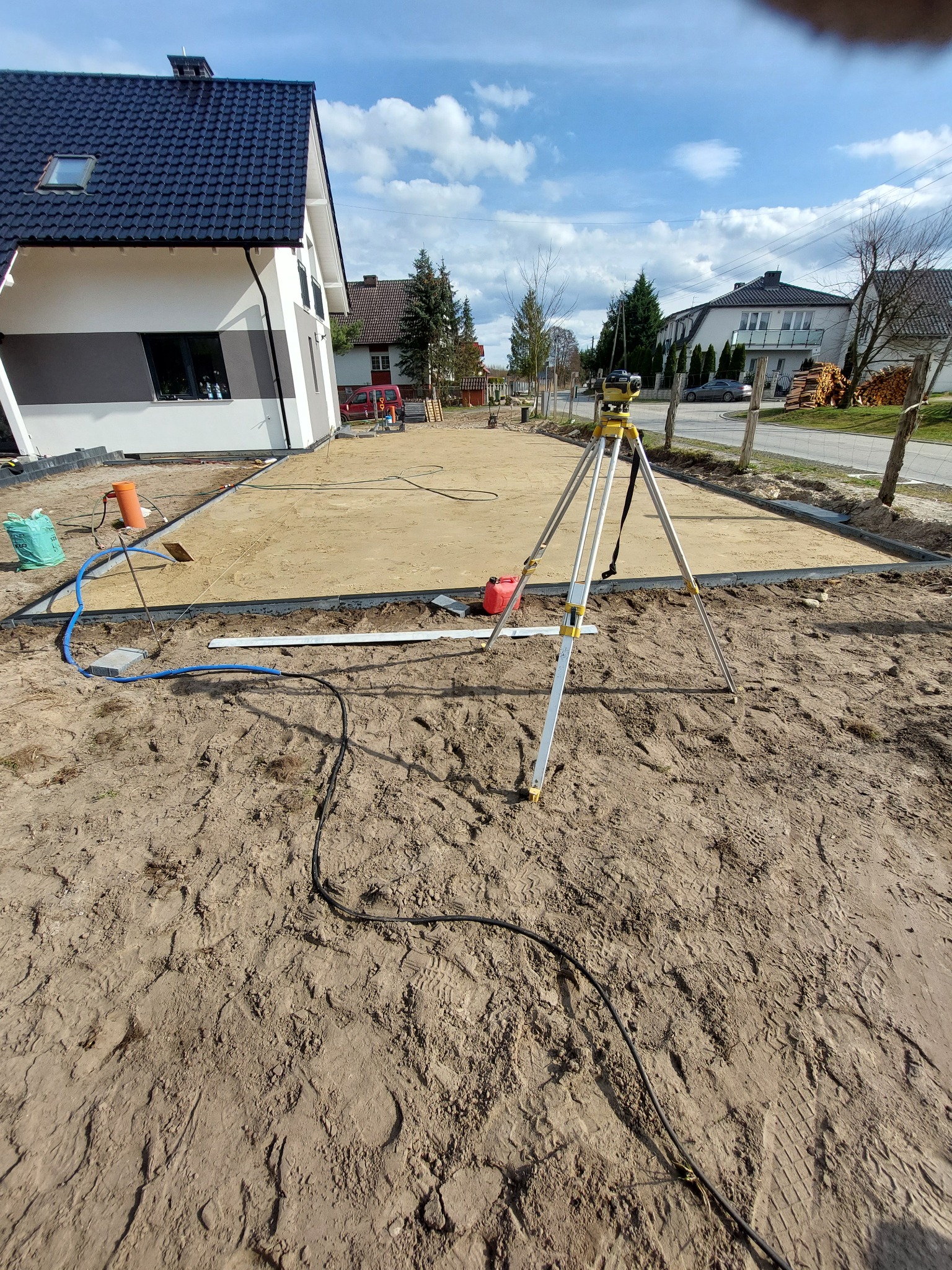 Przygotowany teren pod przyszłą nawierzchnię, z obrzeżami, piaskiem i niwelatorem na trójnogu, widok na dom w tle.