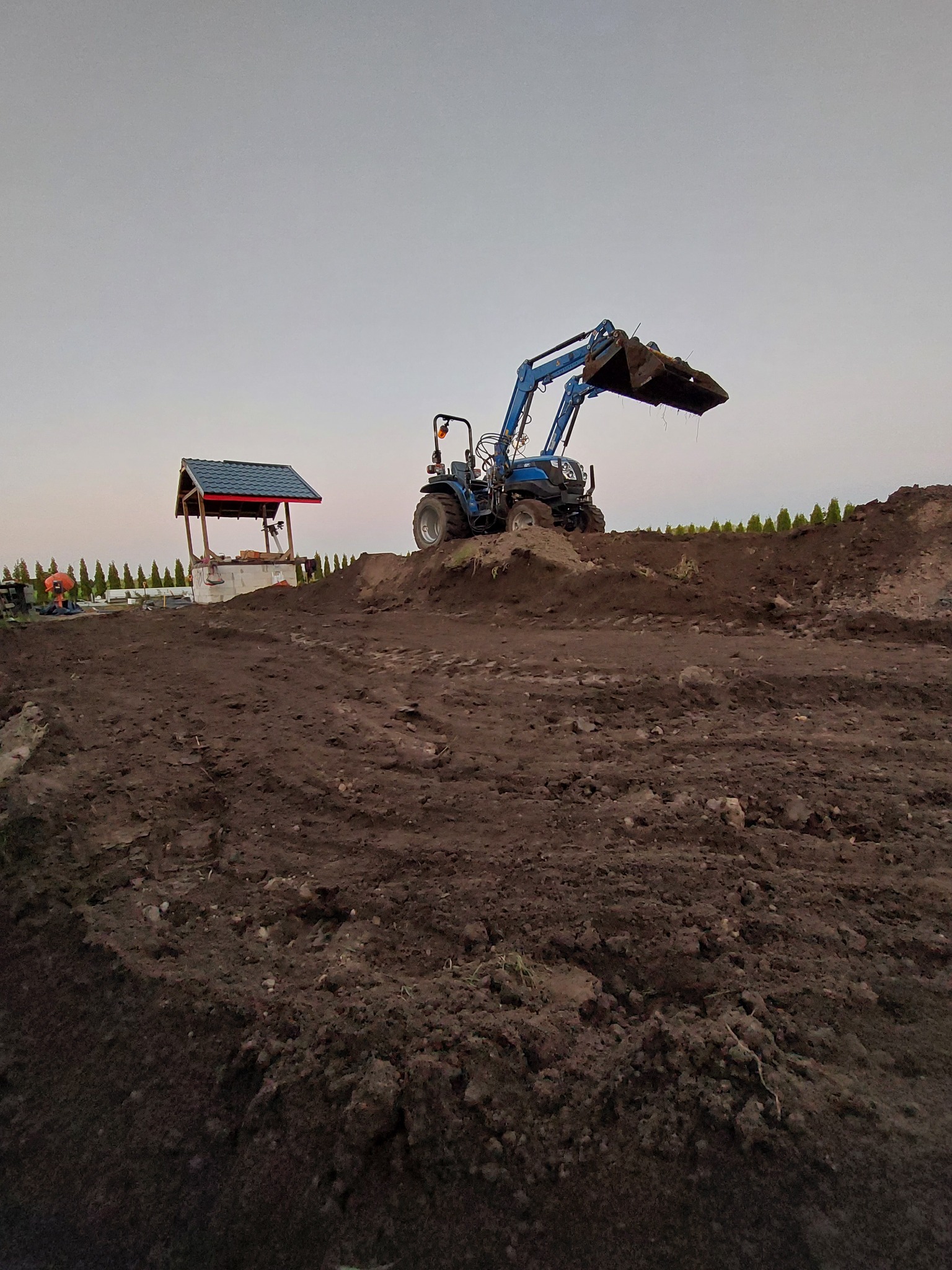 Niebieski traktor z łyżką na kopcu ziemi, w tle studnia z zadaszeniem i rząd zielonych drzewek.