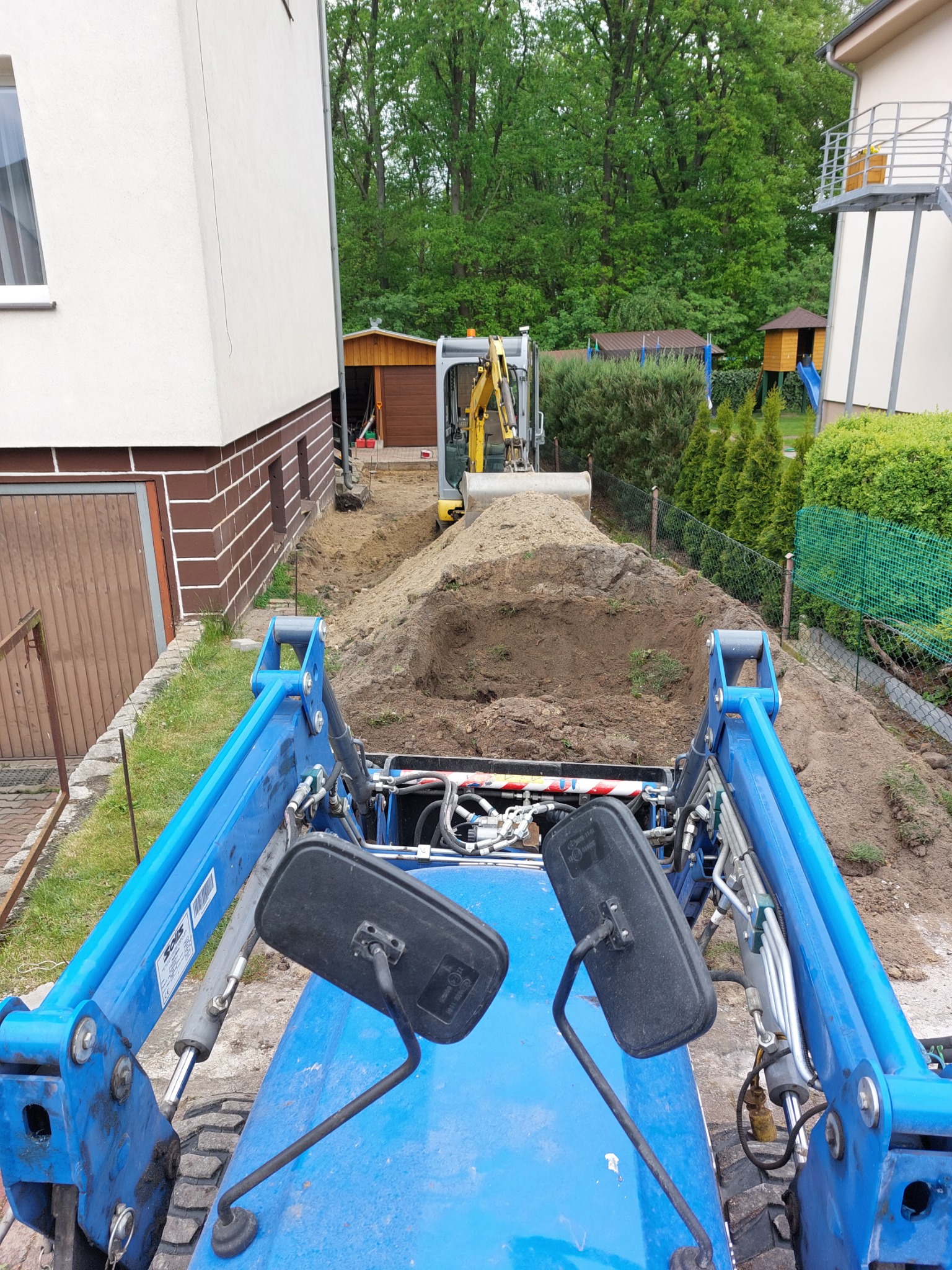 Widok z perspektywy operatora niebieskiego traktora na wykop ziemny z mini koparką w tle, obok domu jednorodzinnego i zieleni.