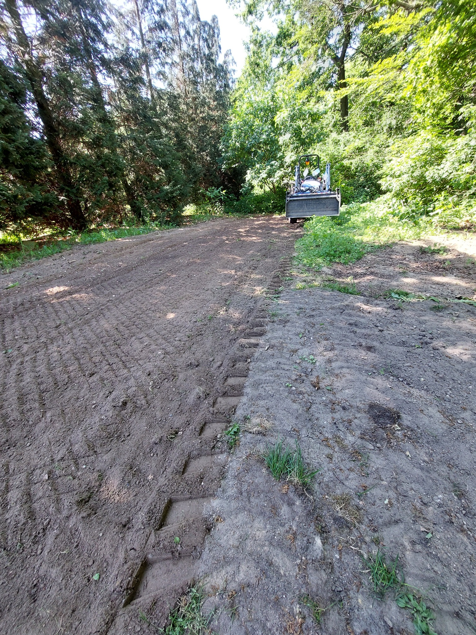 Teren wyrównany traktorem z łyżką, widoczne ślady opon na wilgotnej ziemi, w tle drzewa.