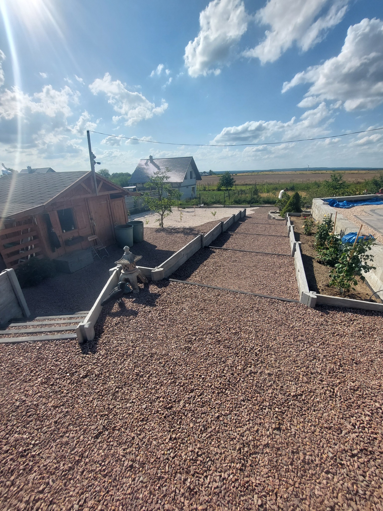 Ogród z ścieżką wysypaną drobnym, czerwonym żwirem, betonowe obrzeża, drewniana altana w tle, widok z góry w słoneczny dzień.