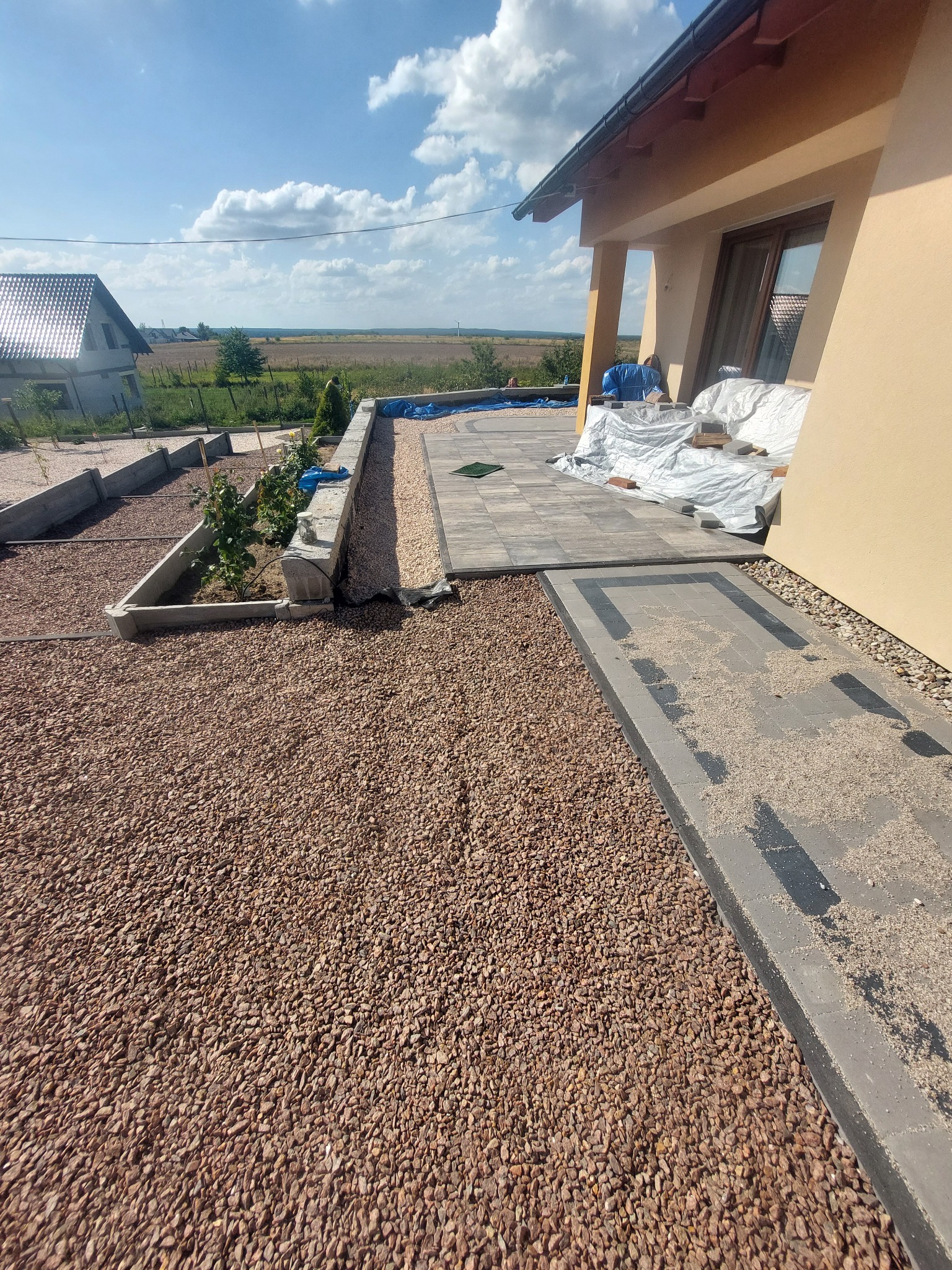 Fragment ogrodu w trakcie prac: ułożona ścieżka z kostki brukowej z ciemnymi wstawkami, otoczona żwirem, widoczne rabaty z roślinami i budynek w tle, częściowo zakryty folią.