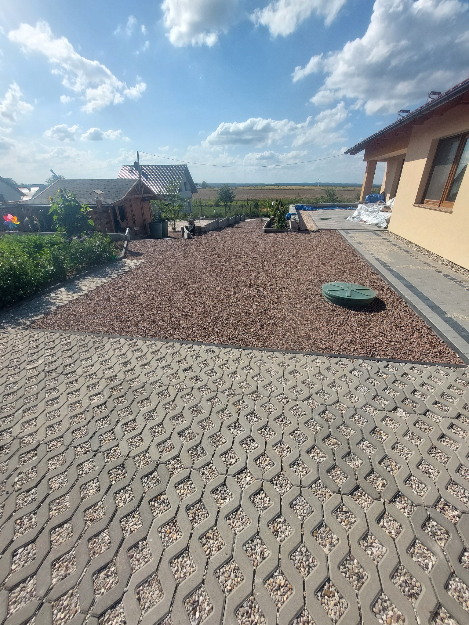 Ułożona szara ażurowa kostka brukowa z wypełnieniem, przechodząca w obszar wysypany drobnym, brązowym żwirem z zieloną pokrywą włazu kanalizacyjnego pośrodku, obok budynku z beżową elewacją...