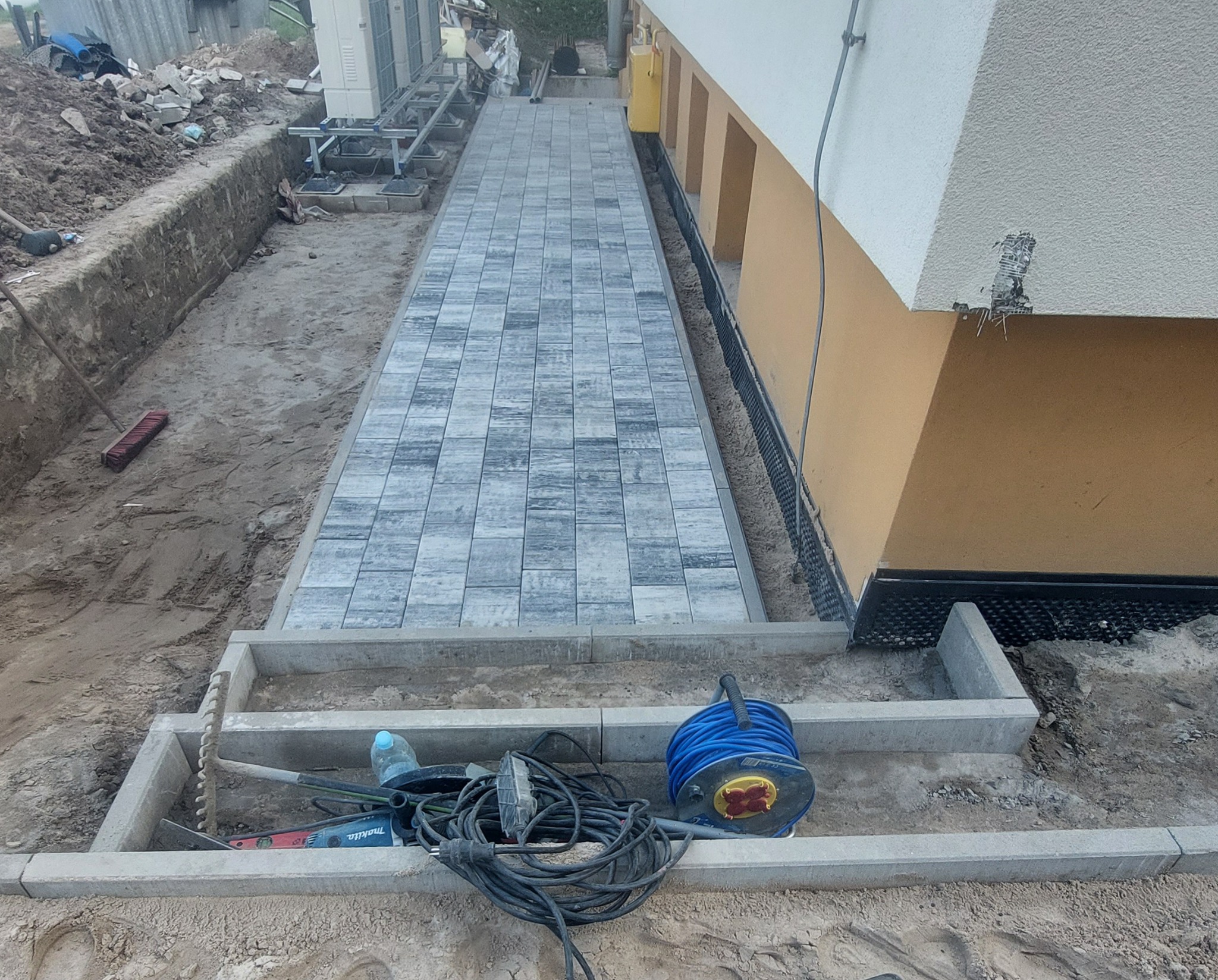 Ułożony chodnik z szarej kostki brukowej prowadzący wzdłuż budynku, widoczne narzędzia i wykop ziemny obok.