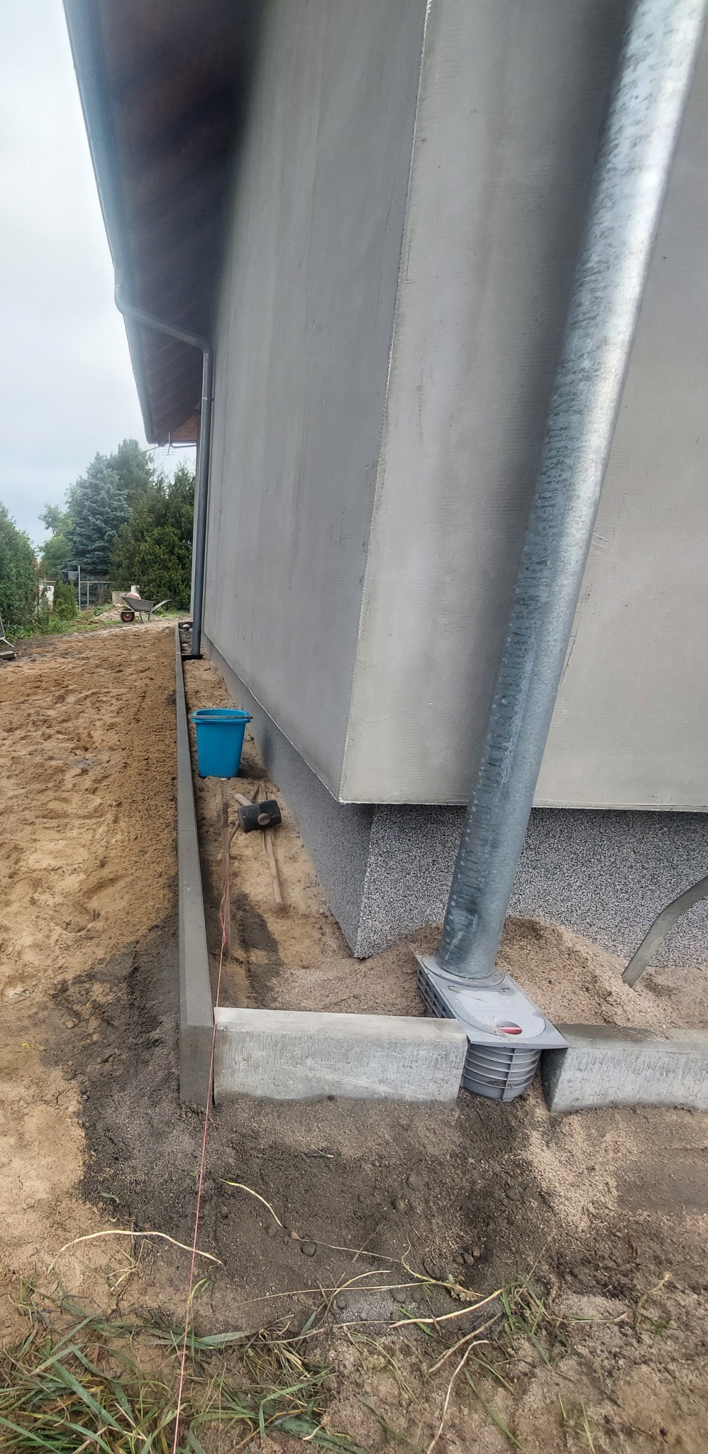 Montaż obrzeży betonowych wokół domu, widoczna linia wyznaczająca poziom, niebieskie wiadro i narzędzia w tle, rura spustowa z systemem odprowadzania wody deszczowej.