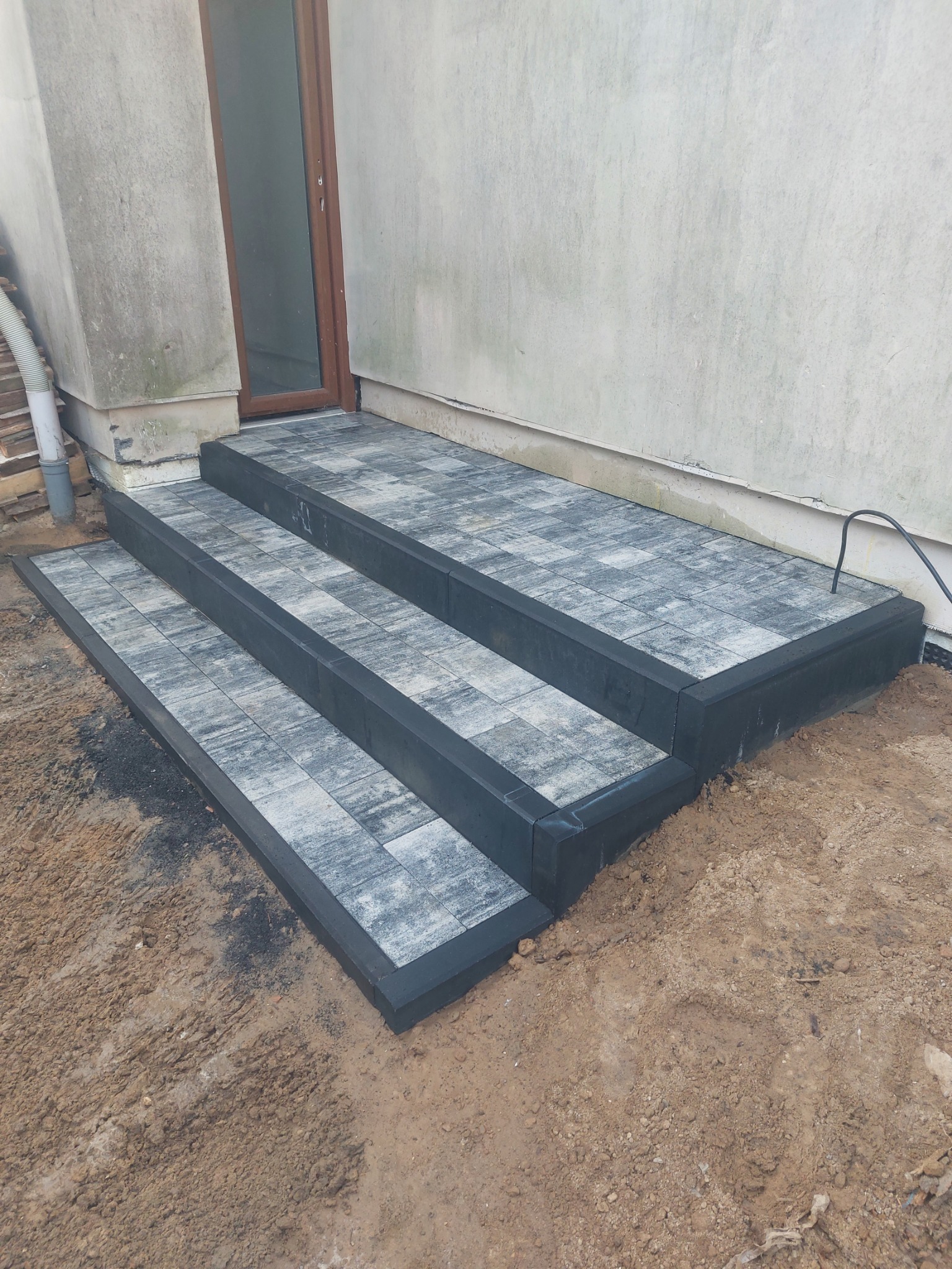 Trzy stopnie schodów wejściowych wykonane z szarej kostki brukowej z czarnymi obrzeżami, prowadzące do brązowych drzwi wejściowych budynku o jasnoszarej elewacji, teren wokół jeszcze...