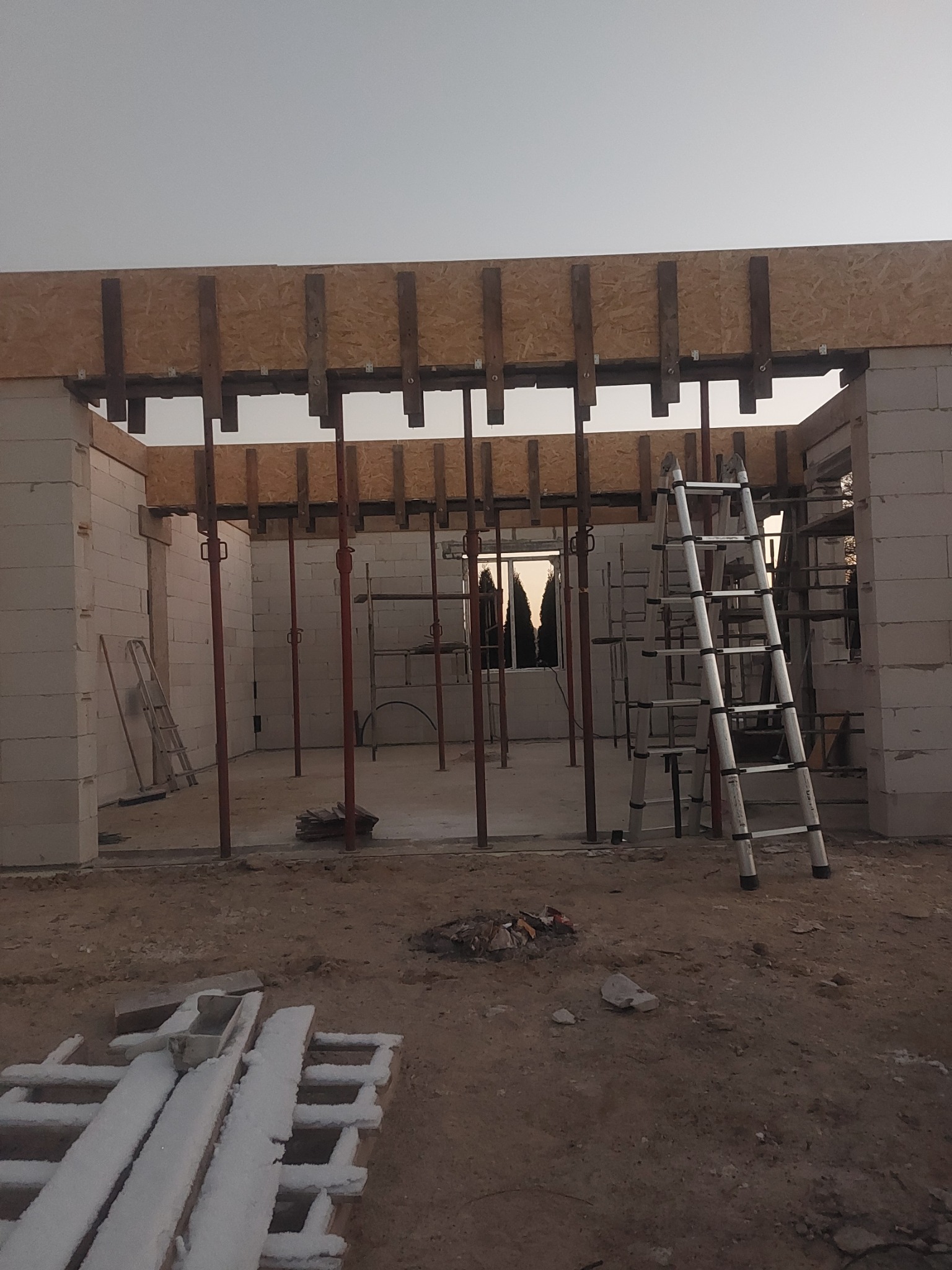 Wnętrze budynku w trakcie budowy z białych bloczków, widoczne stalowe podpory stropu, drewniane szalunki, drabina aluminiowa i resztki śniegu na ziemi.