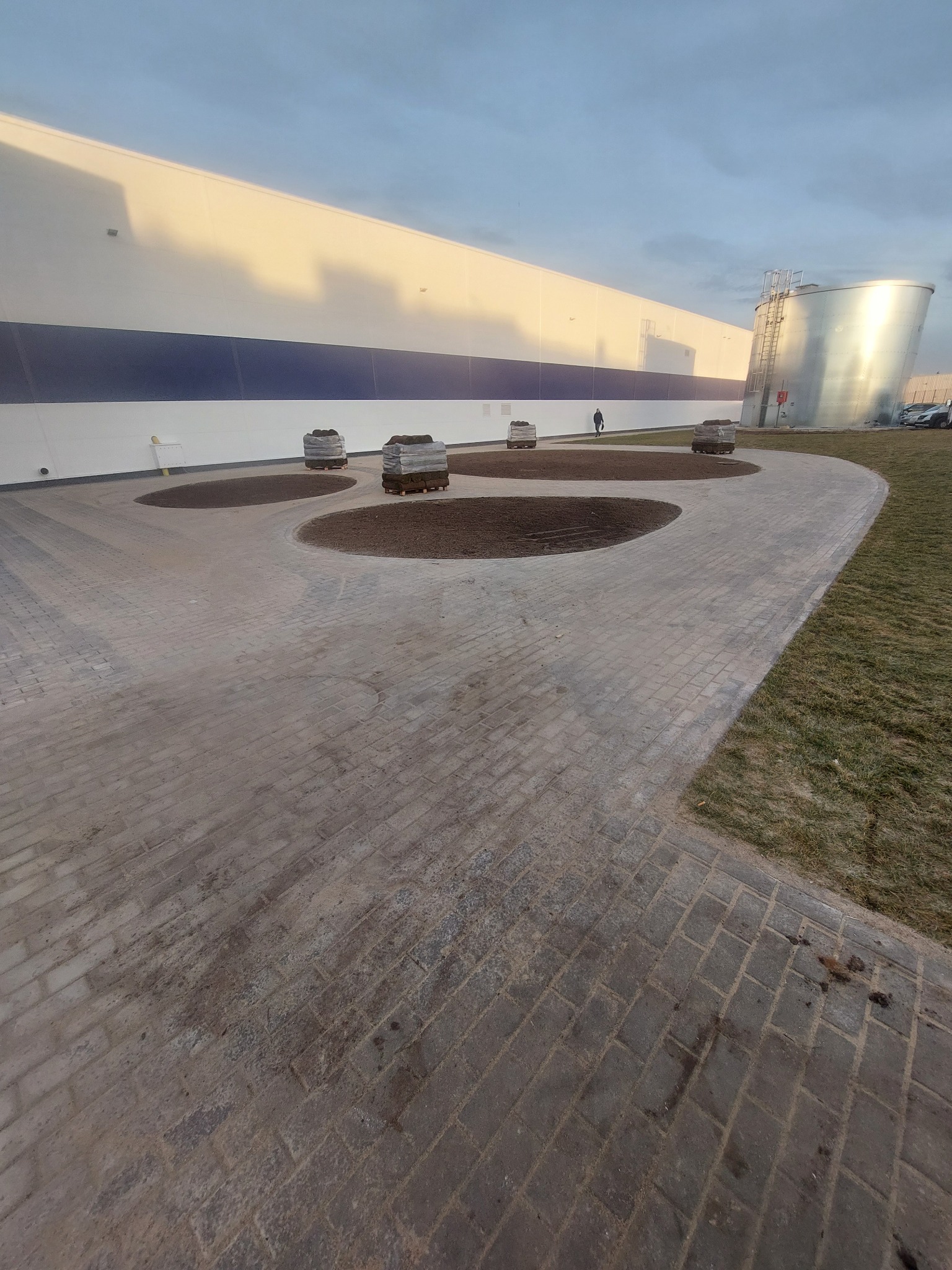 Ułożona kostka brukowa prowadząca do klombów z przygotowaną ziemią pod trawnik z rolki, w tle budynek przemysłowy i zbiornik.